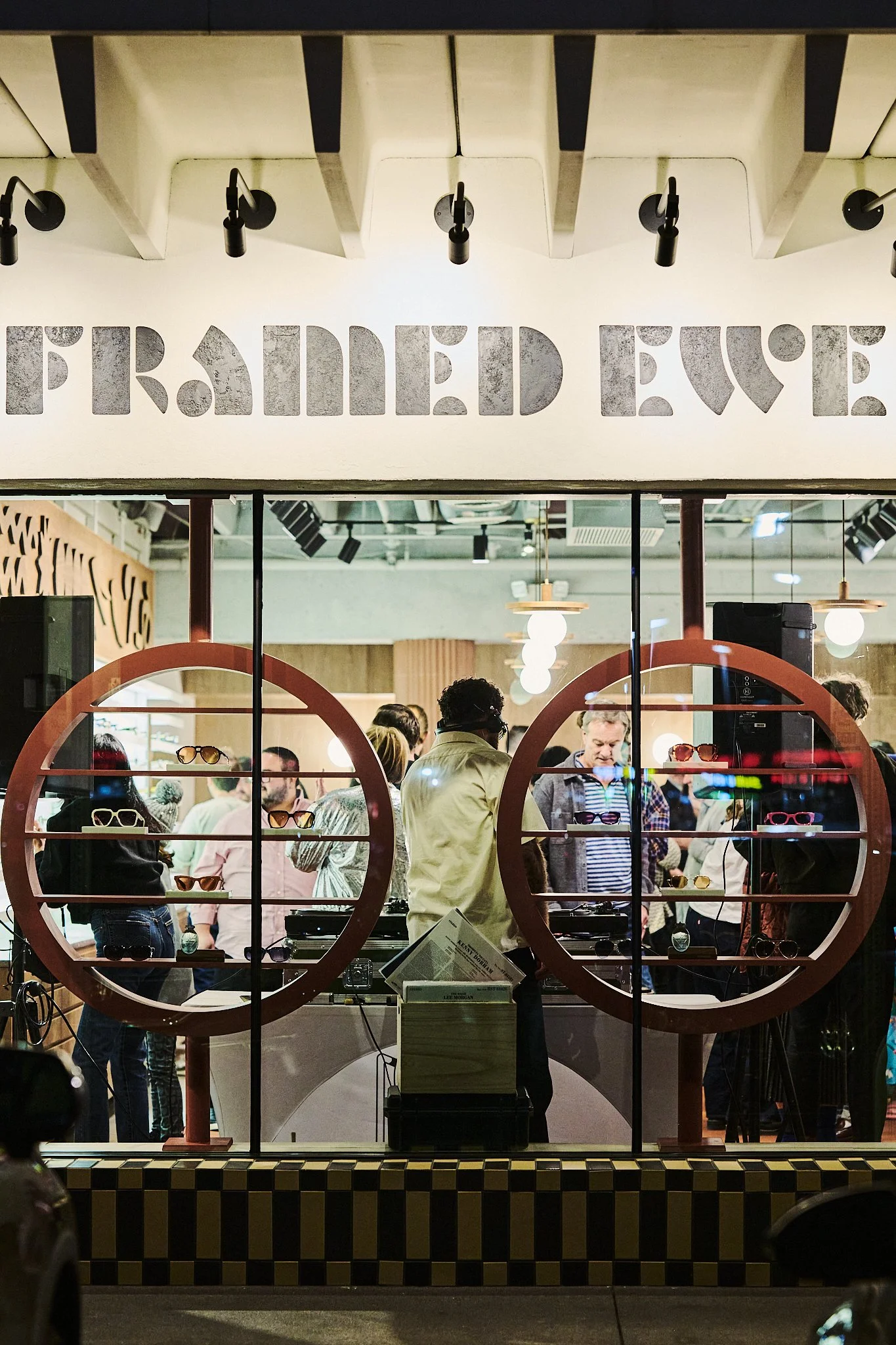 People inside a stylish eyewear store viewed through a window, with sunglasses displayed on round shelves and a sign above reading 'FRAMED EYEWEAR'.