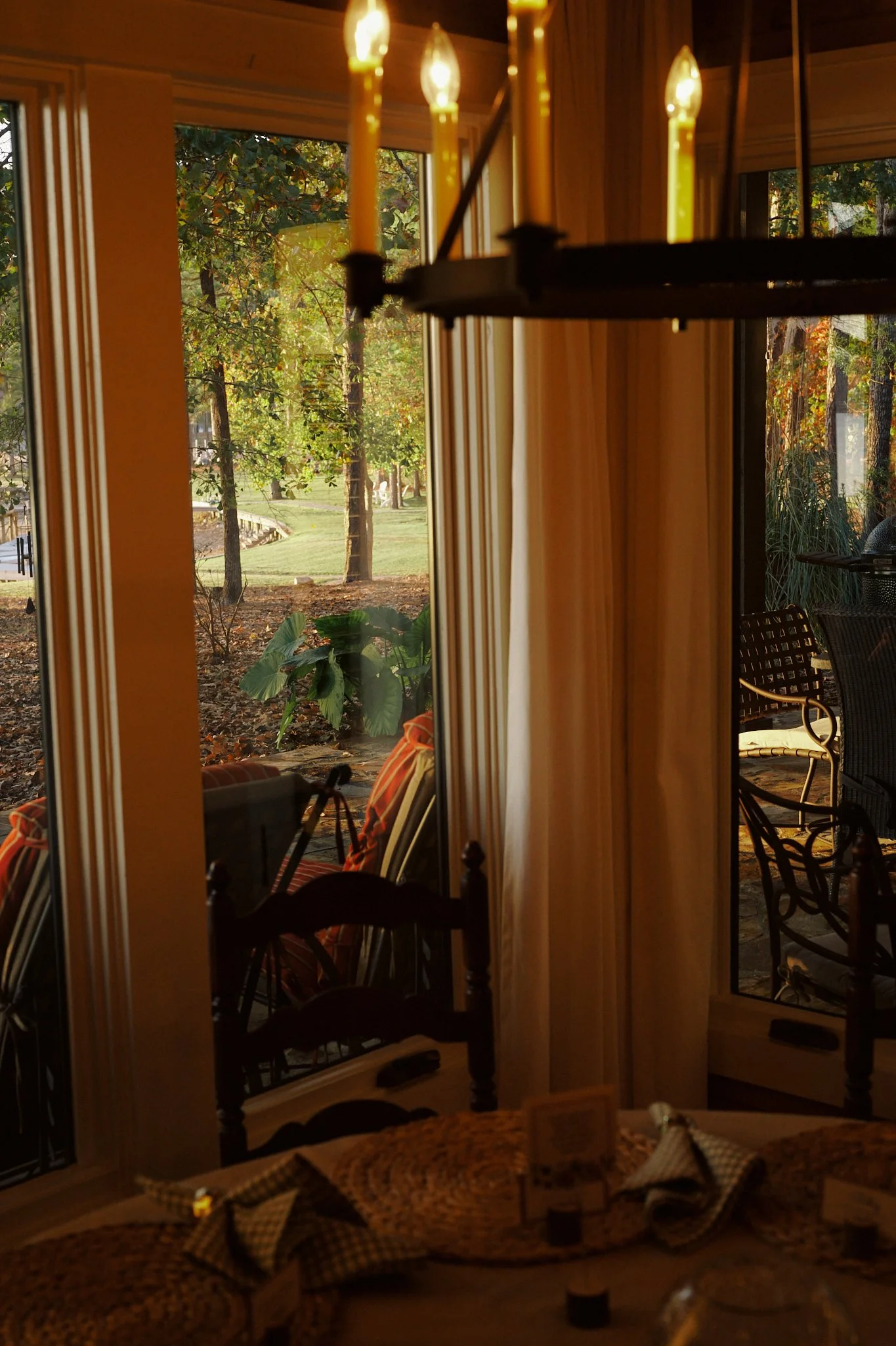 Interior view of a dining room with windows showing a garden with trees and sunlight outside, and a chandelier with lit candles hanging from the ceiling.
