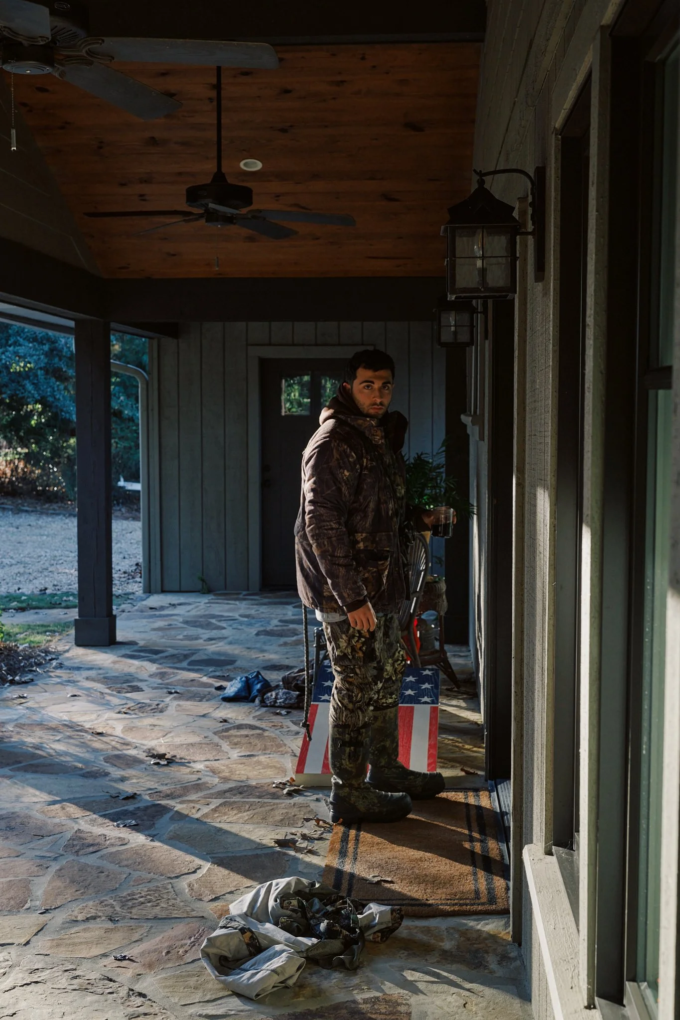 A man standing on a stone porch holding a drink, wearing camouflage clothing, with a door behind him and outdoor foliage visible.