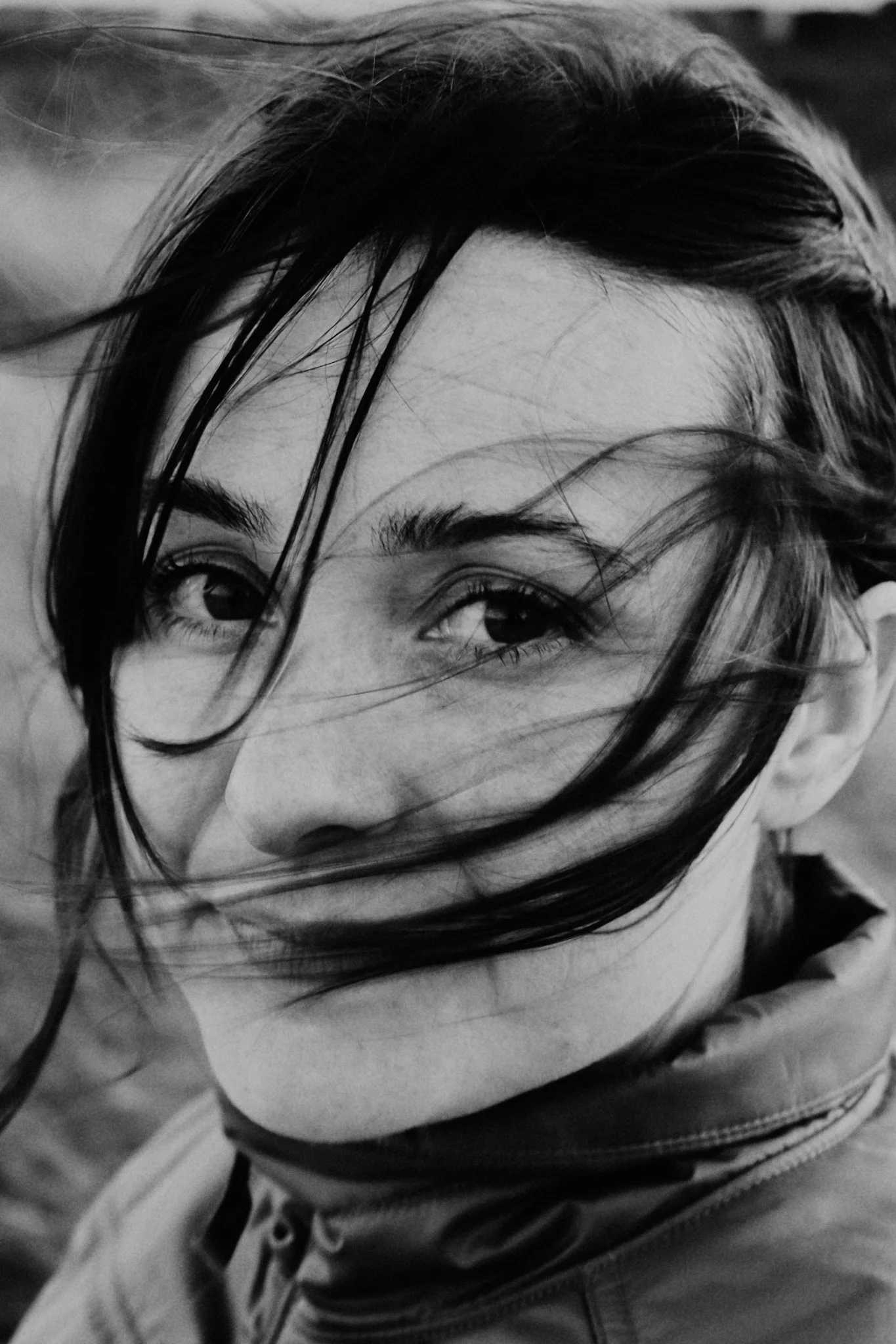Close-up black and white portrait of a woman with wet hair covering part of her face, smiling softly, wearing a jacket.