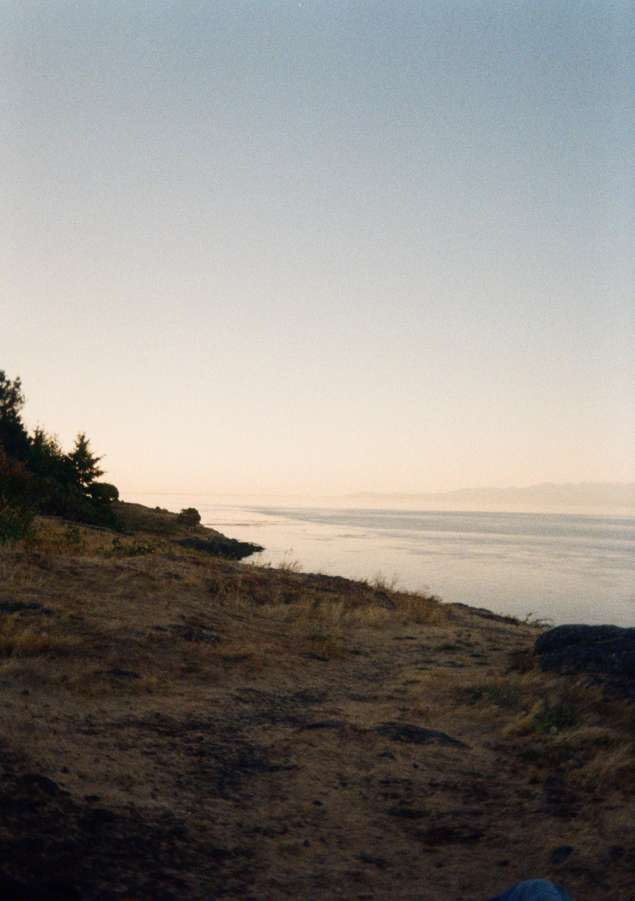 A coastal landscape during sunset with a rocky beach, sparse vegetation, and calm ocean waters extending to the horizon under a clear sky.
