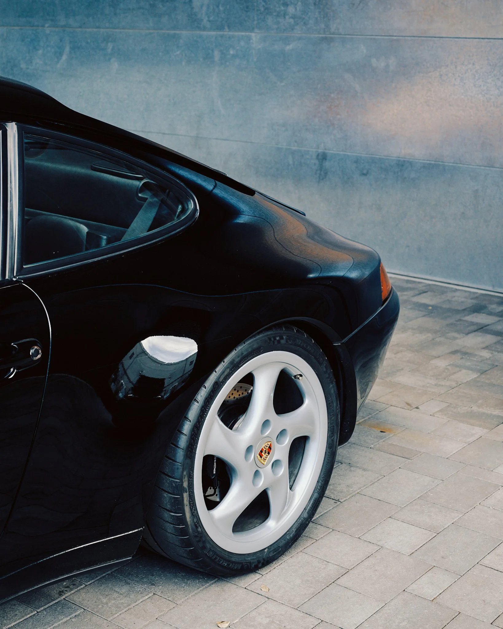Close-up of the rear side of a black Porsche sports car, showing the white alloy wheel with the Porsche crest, parked on a paved surface against a textured gray wall.