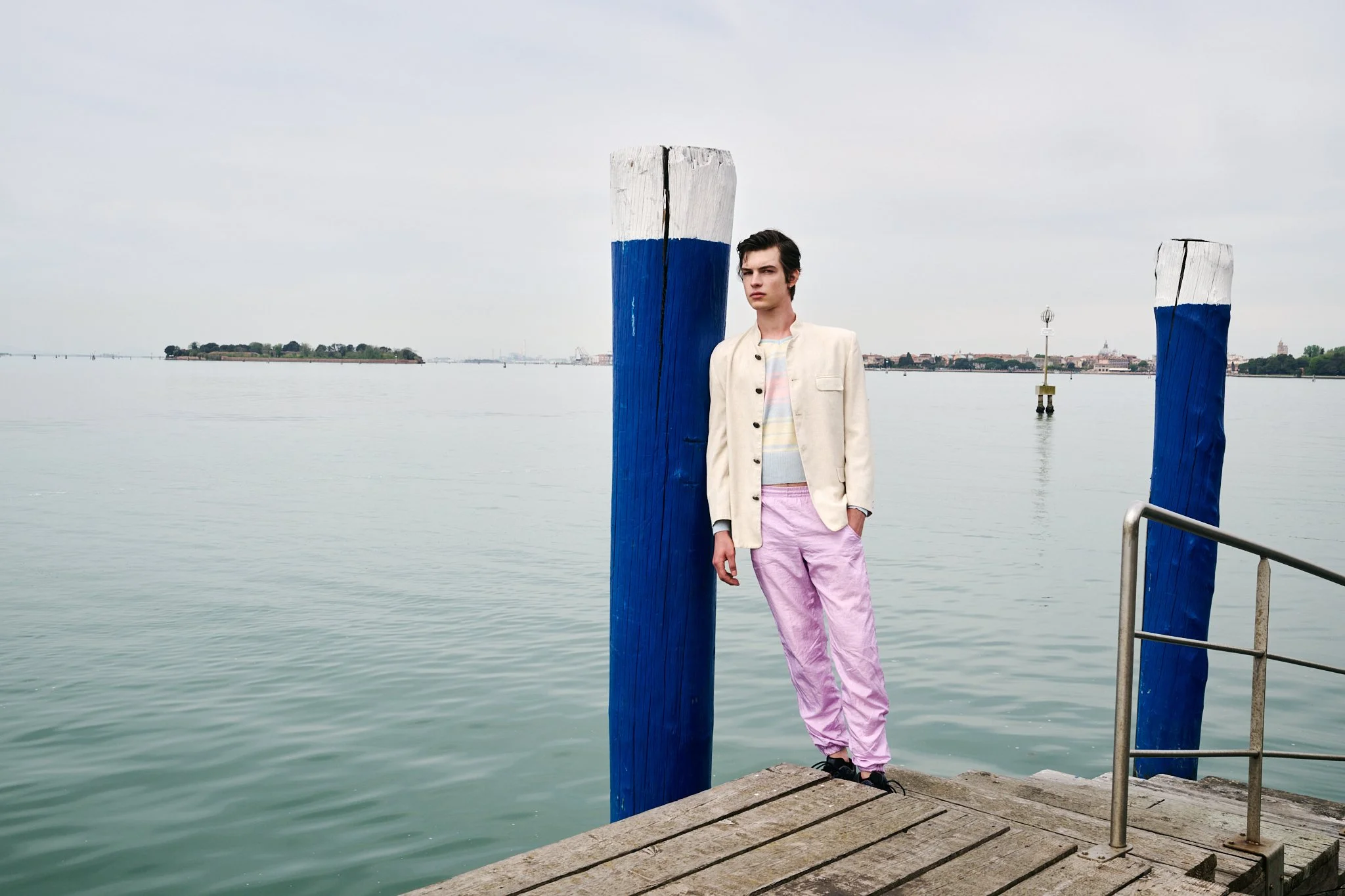 A young man with dark hair standing on a wooden dock by the water, wearing pastel pink pants, a light-colored jacket, and a striped shirt, with a cloudy sky in the background.