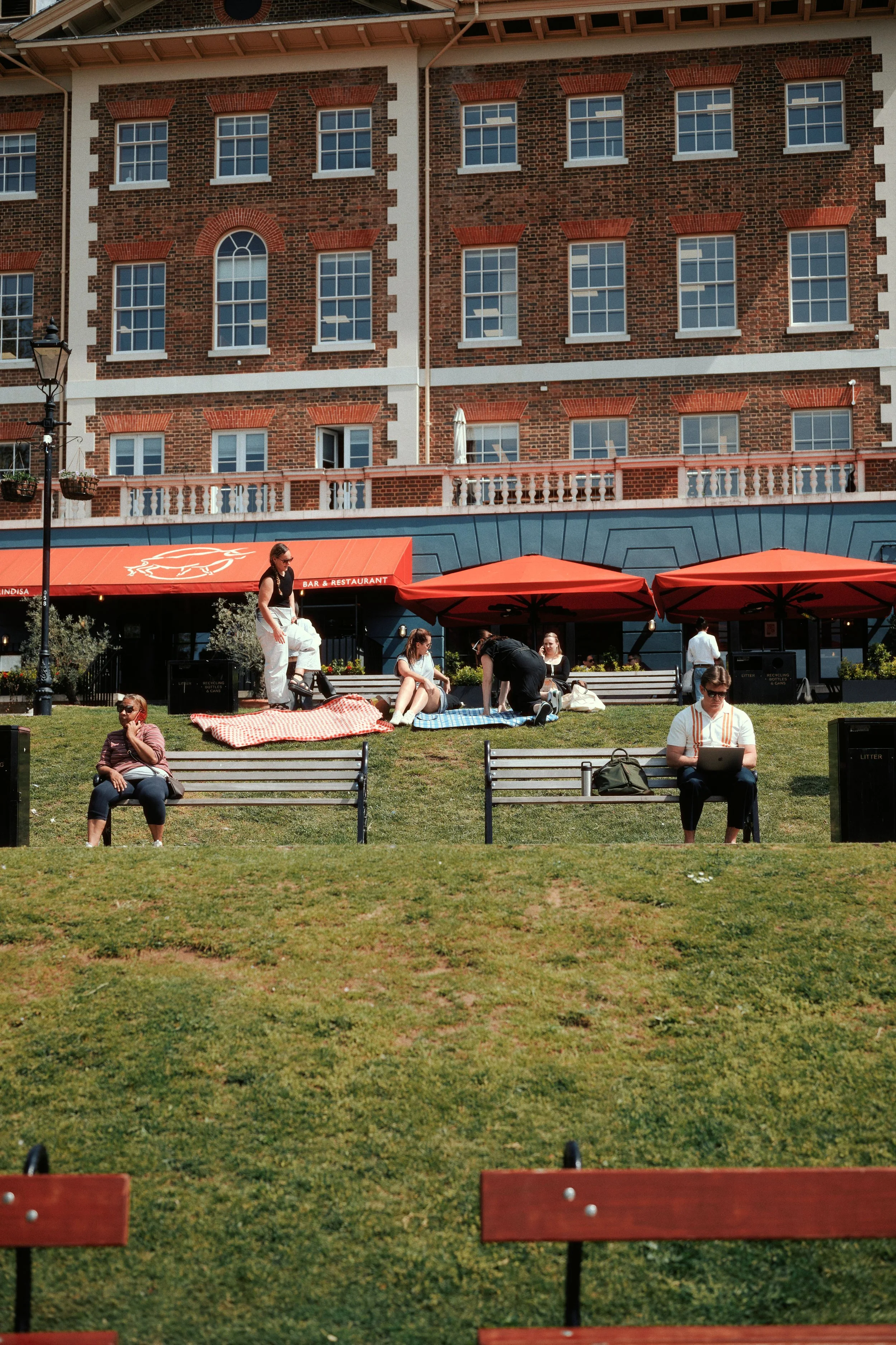 People relaxing and socializing on a grassy park area outside a building with red brick facade, with outdoor seating, umbrellas, and benches.