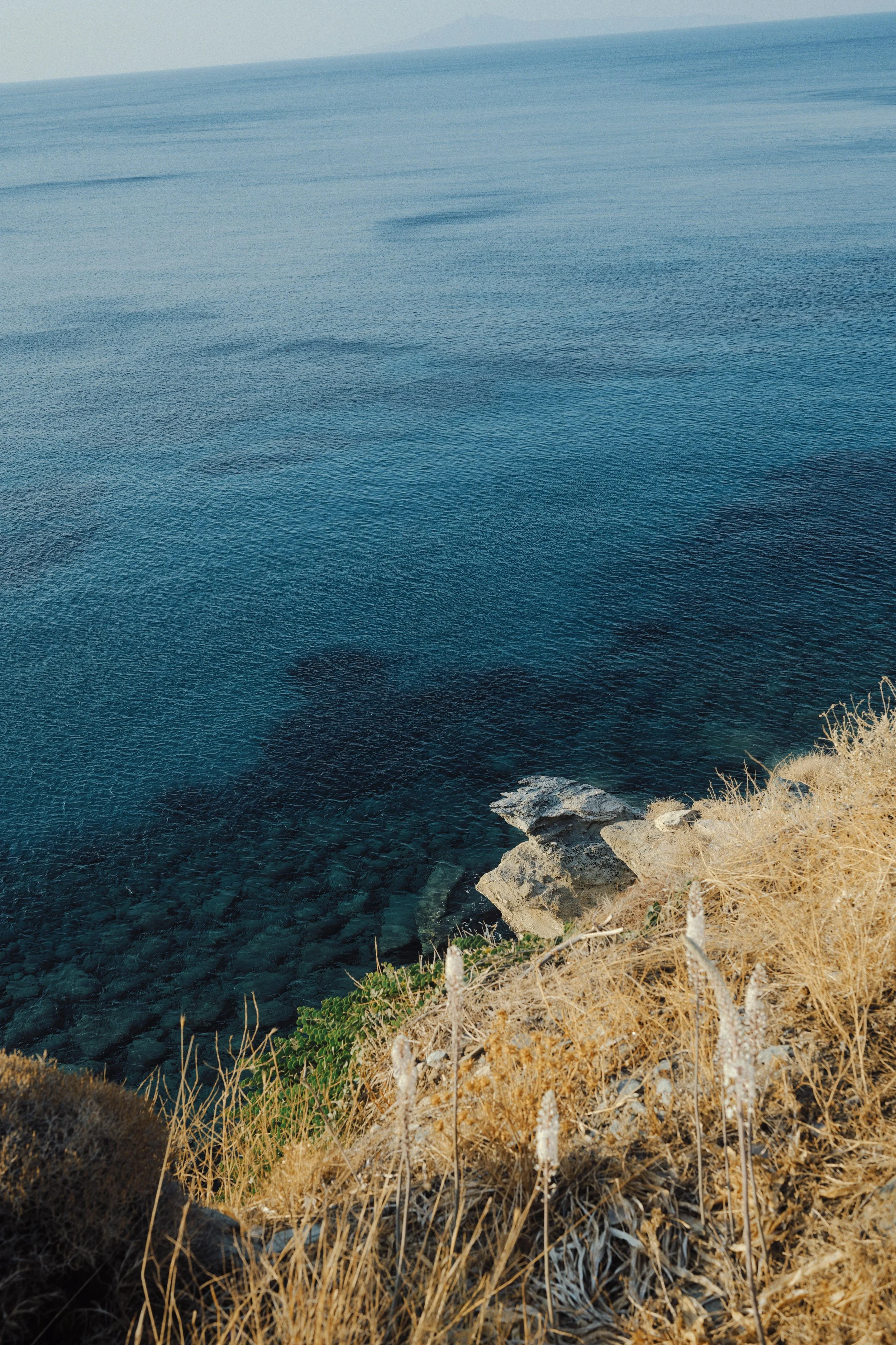 Cliffside view of the ocean with clear water and rocky shoreline, dry grass and sparse vegetation in the foreground.