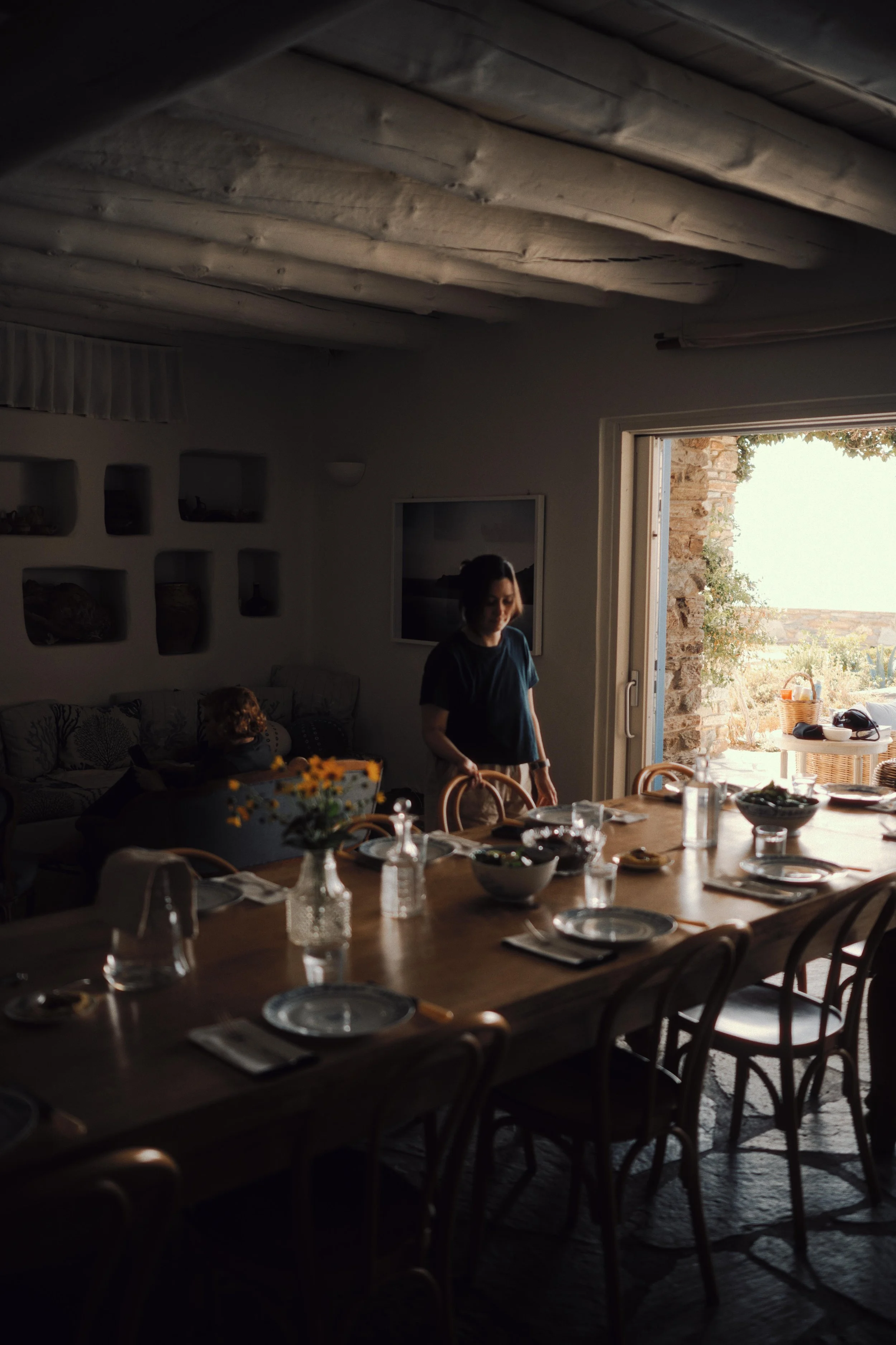 A woman is setting the dining table in a cozy, rustic room with natural light from an open door, a long wooden table with plates, glasses, and bowls, and a woman sitting in the background.