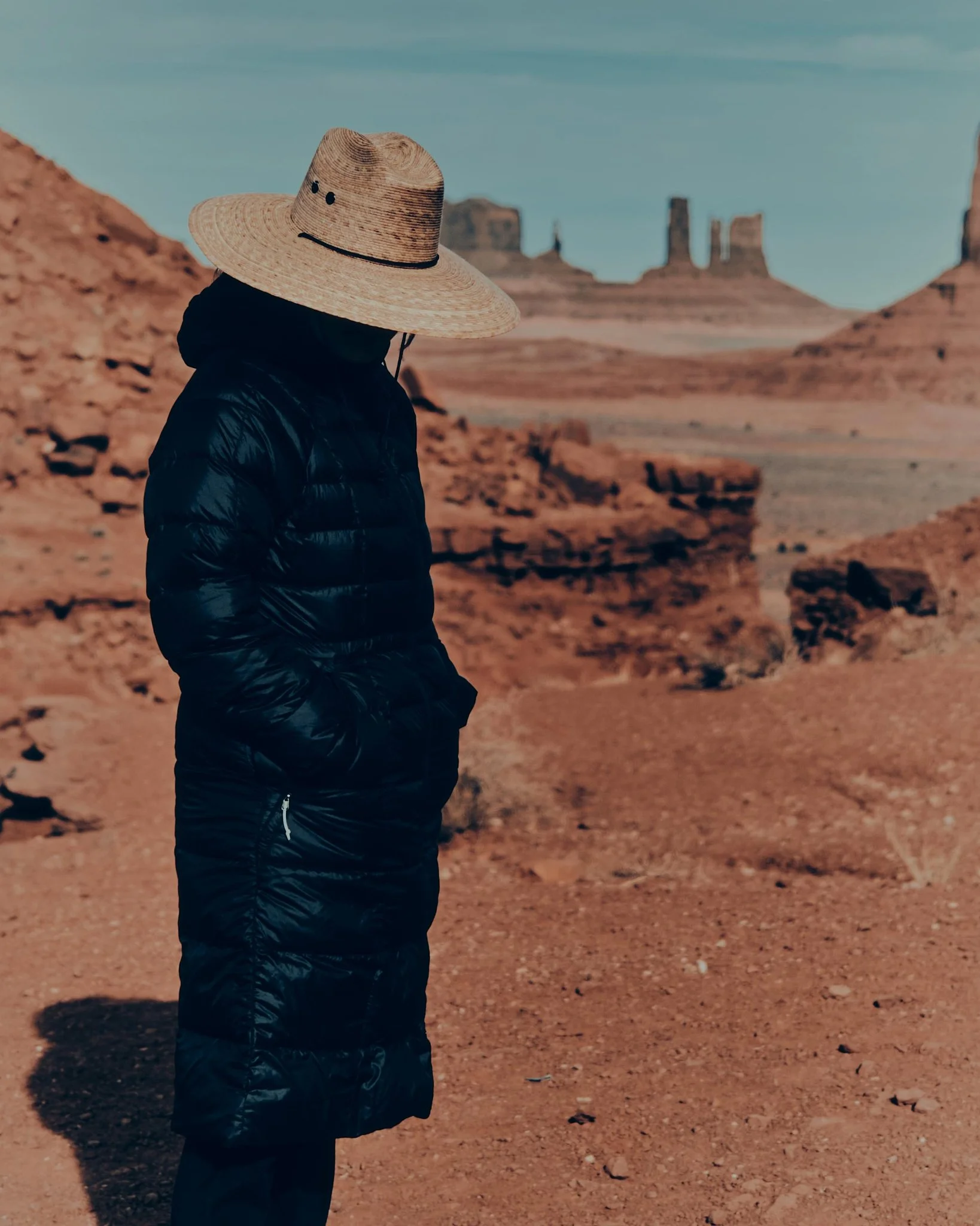 Person wearing a large straw hat with black dots and a smiley face, black puffy jacket, standing in a desert landscape with orange rocks and rock formations in the background.