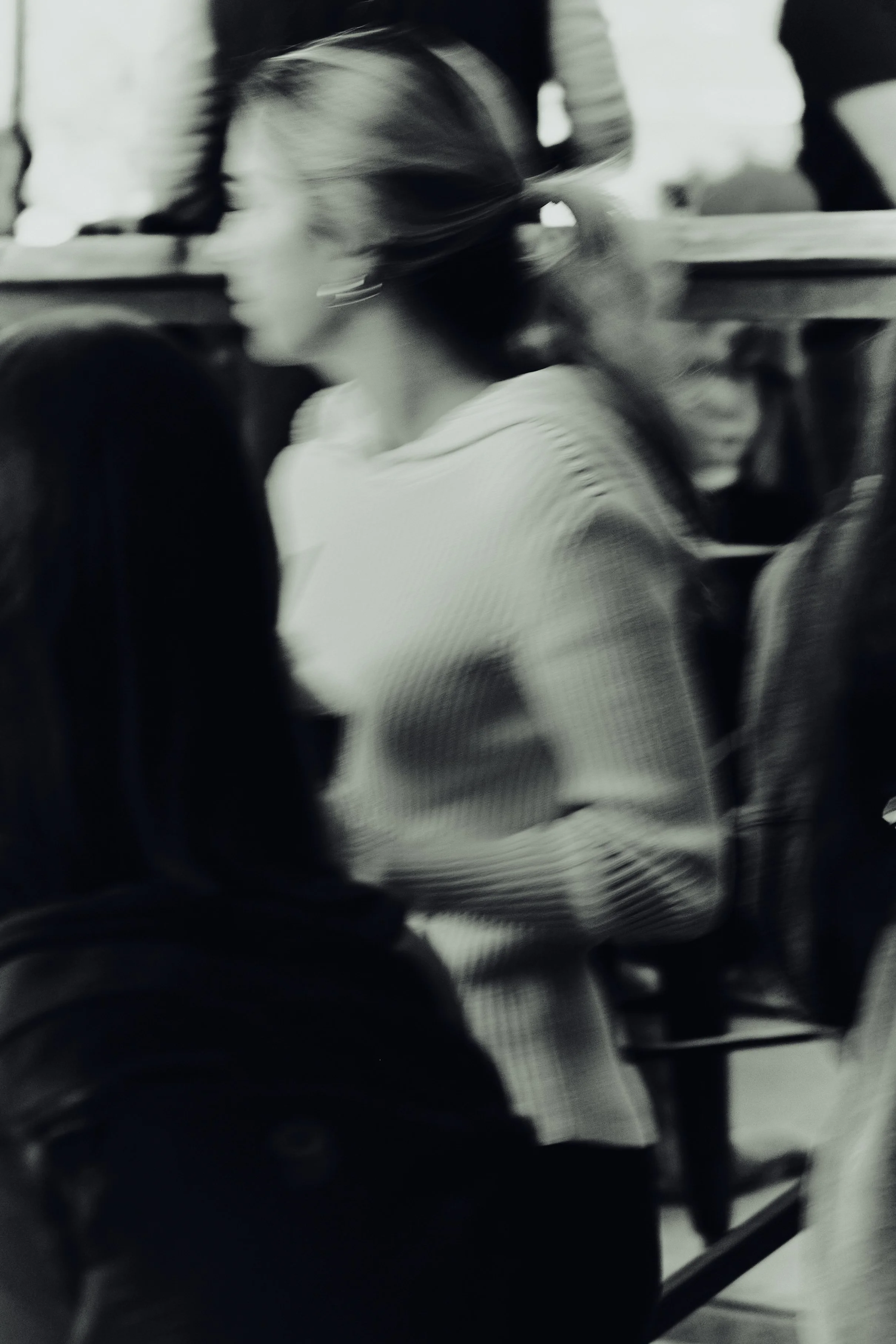 Blurred black-and-white photo of a woman with glasses, sitting in a crowded space, possibly a train or bus, facing to the side.