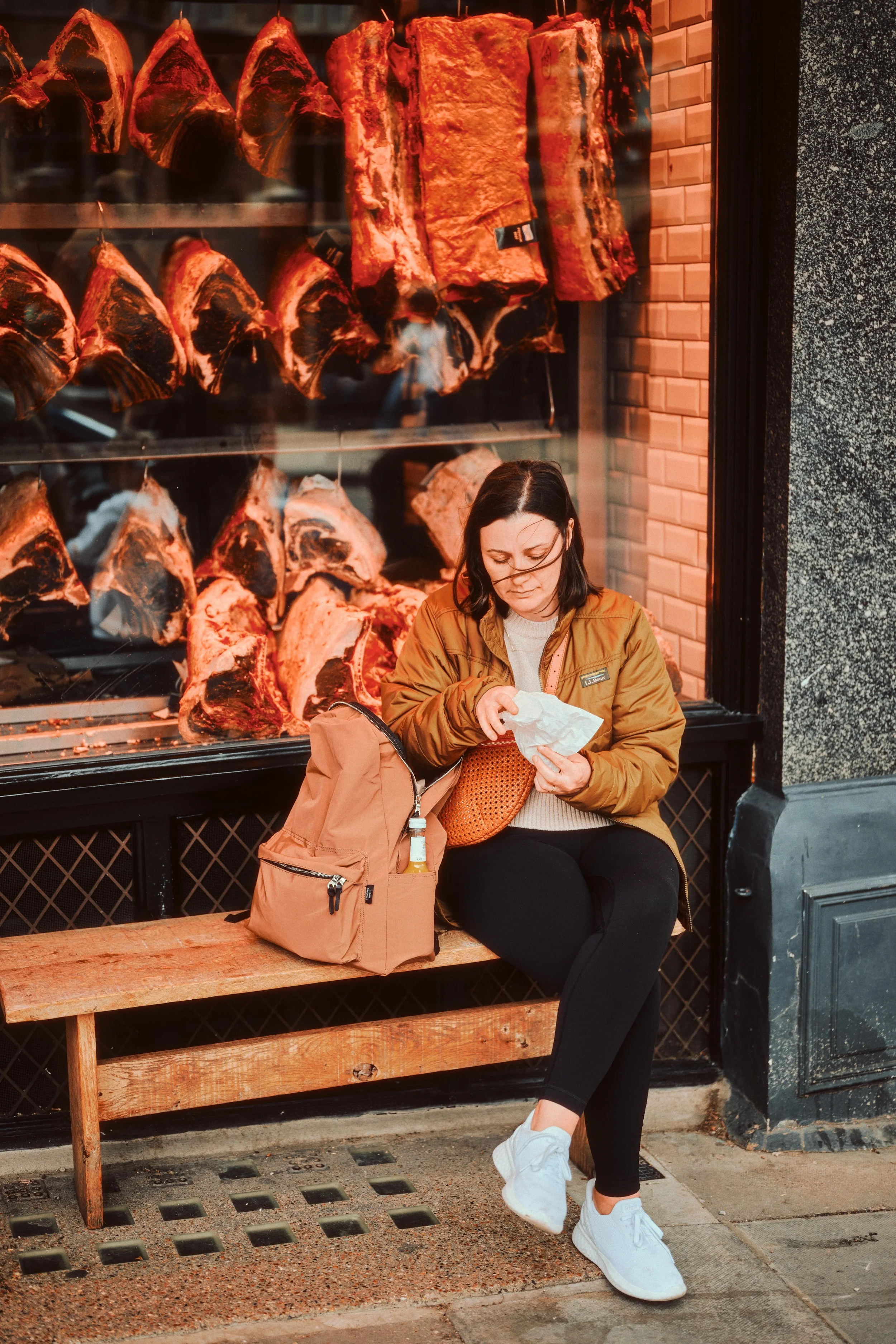 A woman sitting on a wooden bench outside a butcher shop with raw meat slabs displayed behind her, looking into a paper bag.