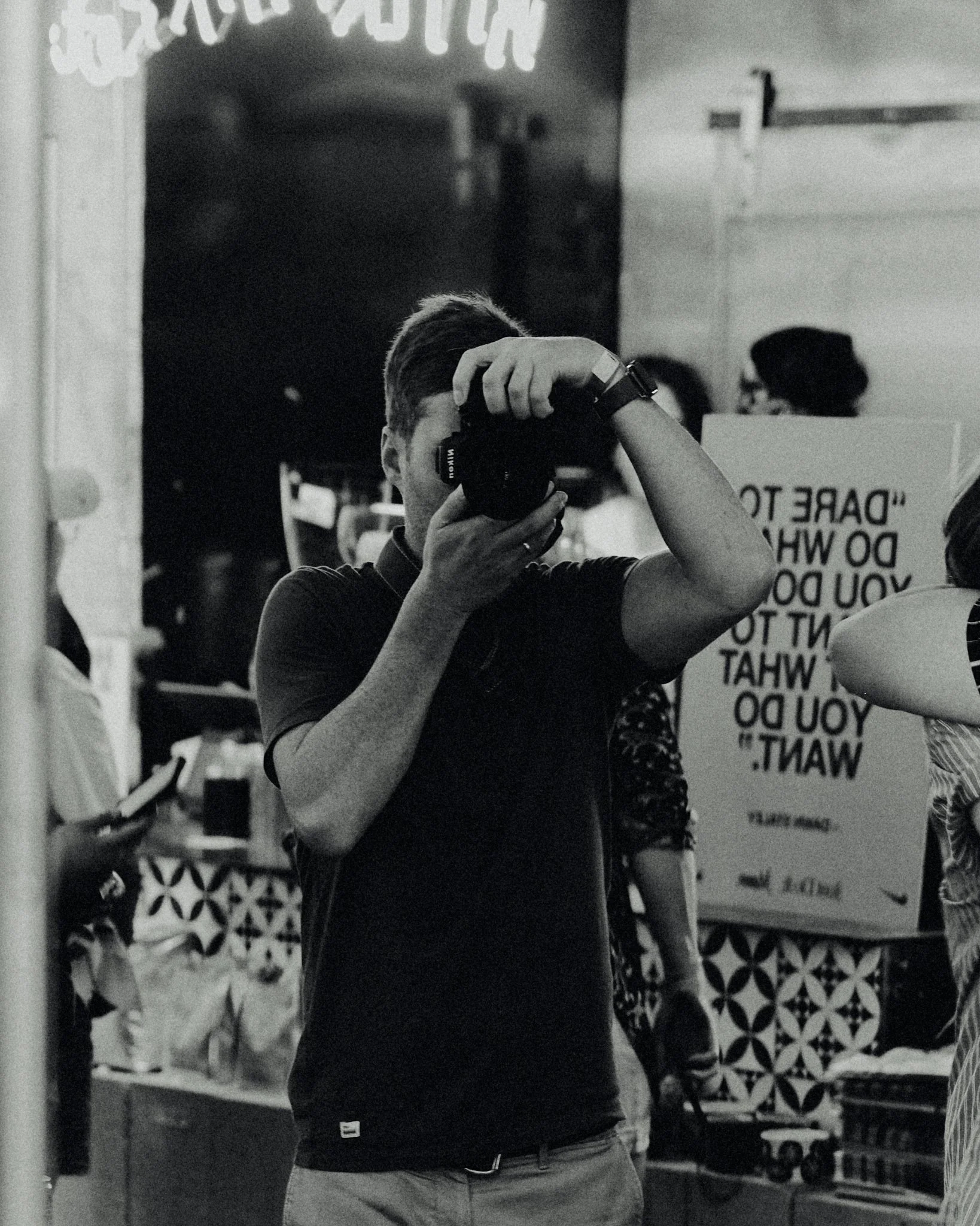 A person taking a photo with a camera in a room filled with people and display signs, in black and white.