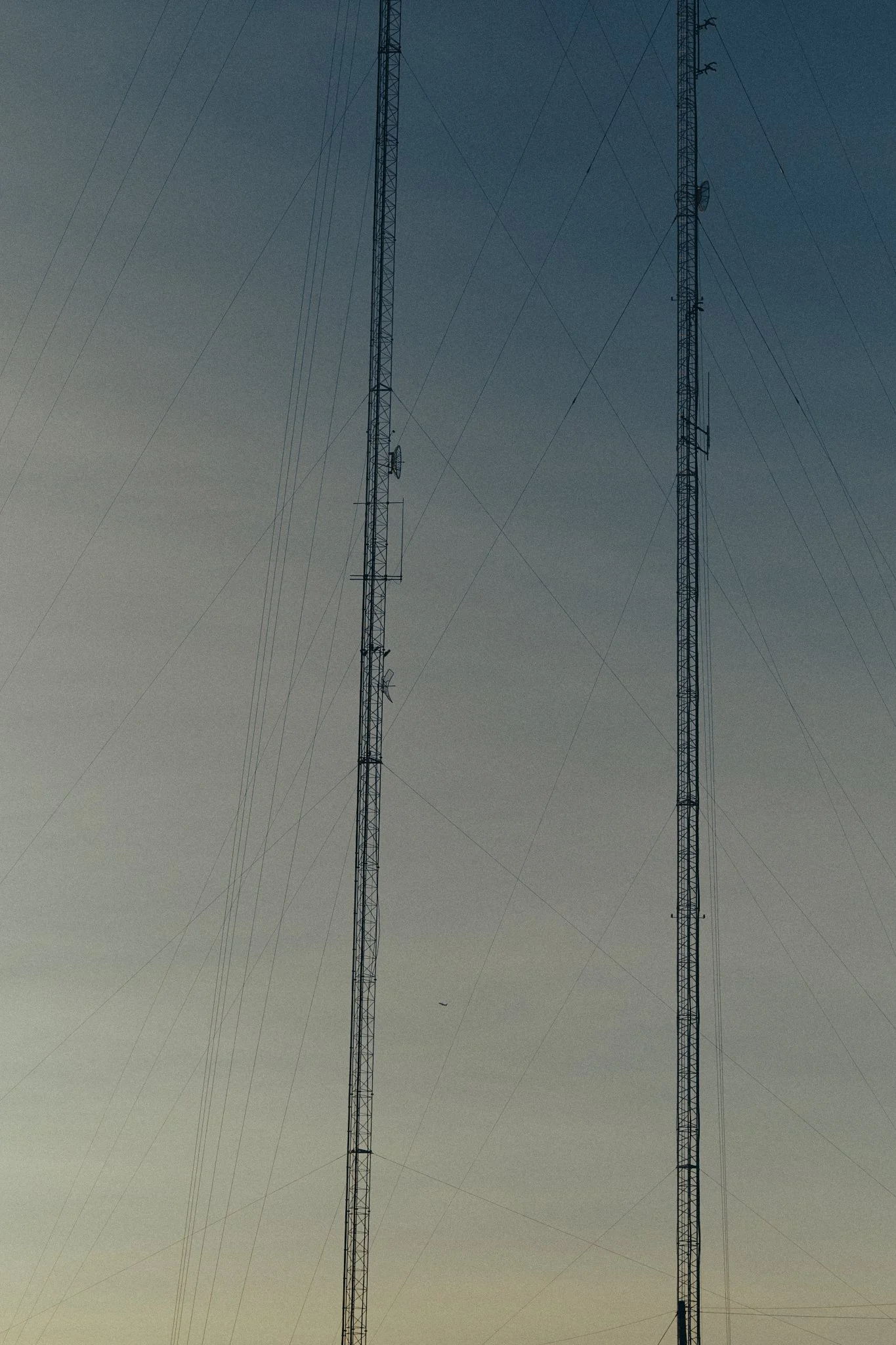 Two tall radio towers with multiple antennae against a gradient sky, with a small airplane visible in the distance.