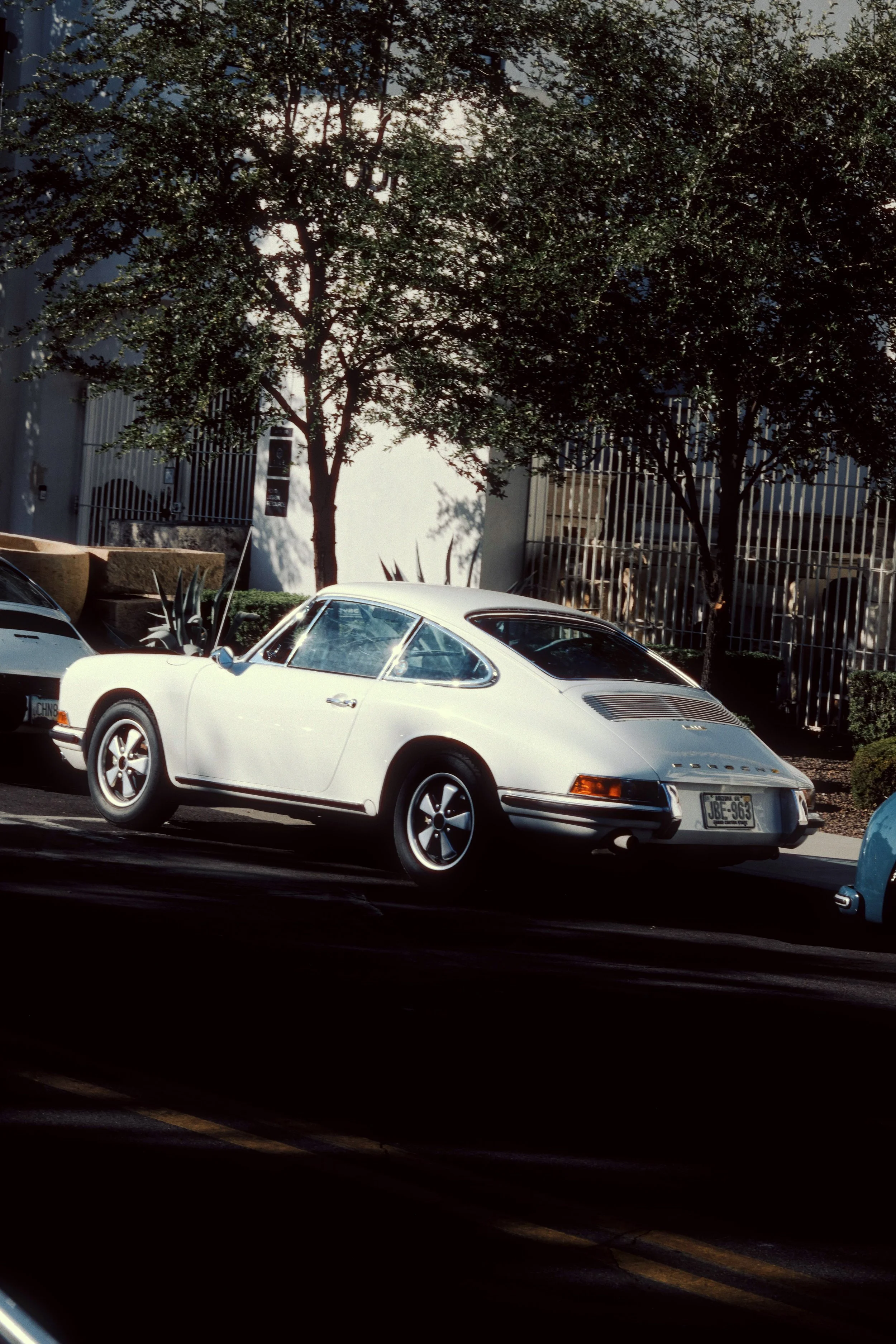 A white classic Porsche 911 parked on a street, with trees and buildings in the background.