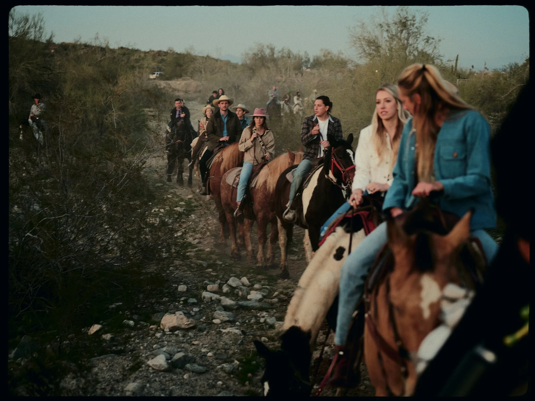 A group of people riding horses along a rocky dirt trail in a desert landscape. Some are dressed in casual western attire, with a few wearing hats.
