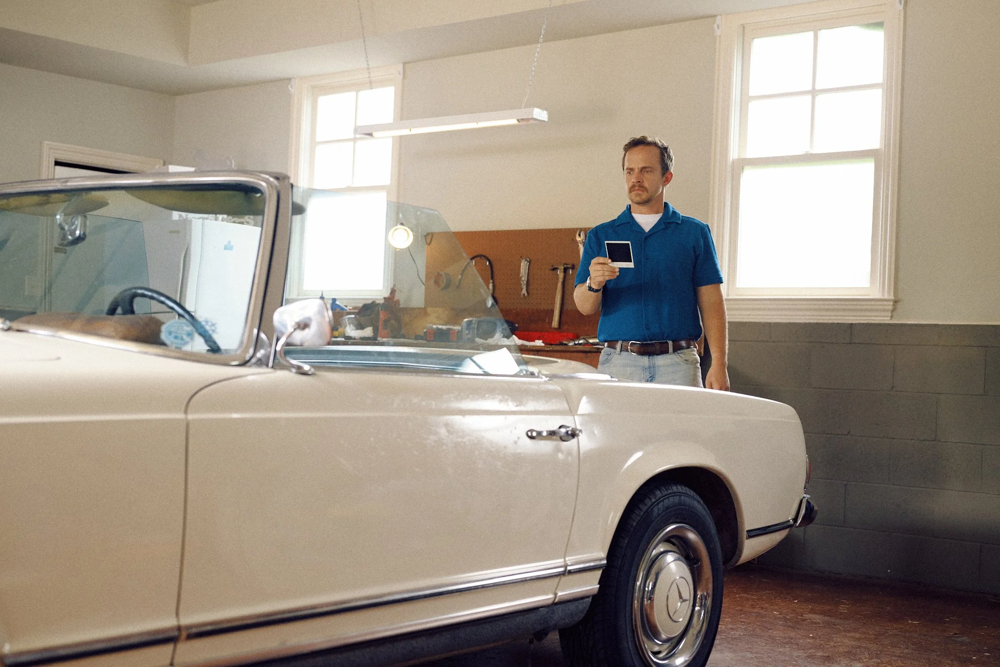 A man standing in a garage, looking at a photo or screen, with a vintage beige Mercedes-Benz convertible car in the foreground and tools on a workbench in the background.
