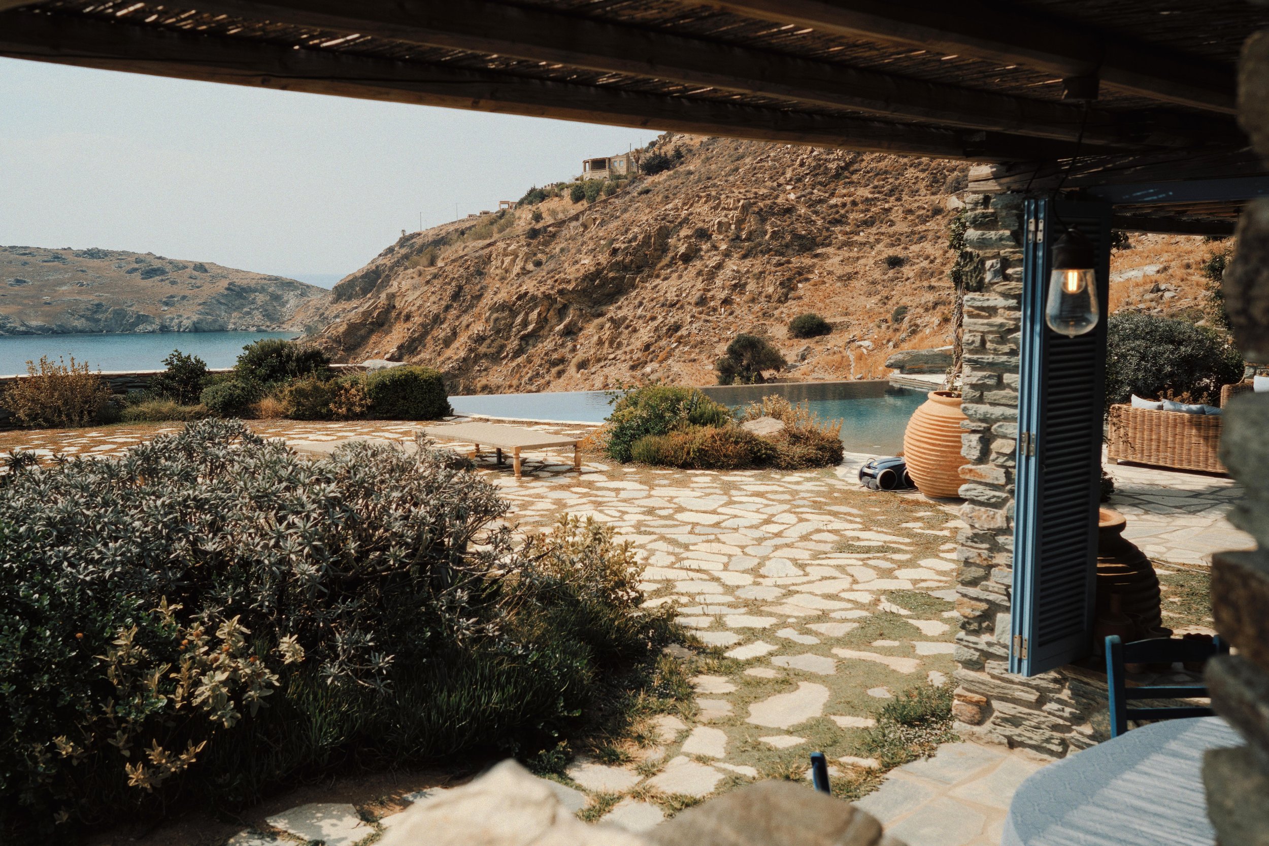 View of a relaxed outdoor patio overlooking a hill with dry vegetation and a body of water, with a stone and wood structure, a hanging light bulb, a large pottery jar, and outdoor seating.