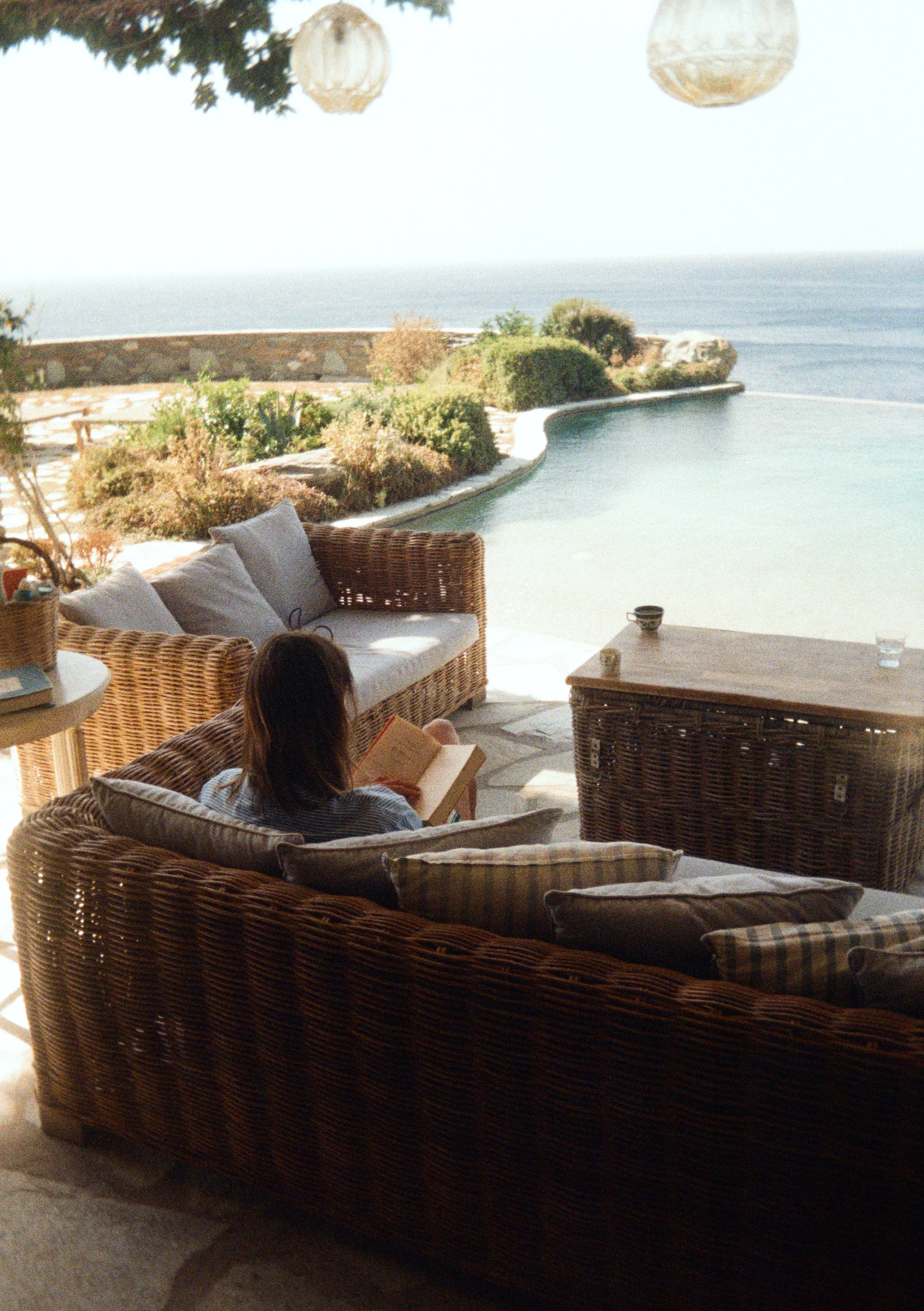 A person relaxing on a wicker sofa, reading a book, with a garden and swimming pool overlooking the ocean in the background.