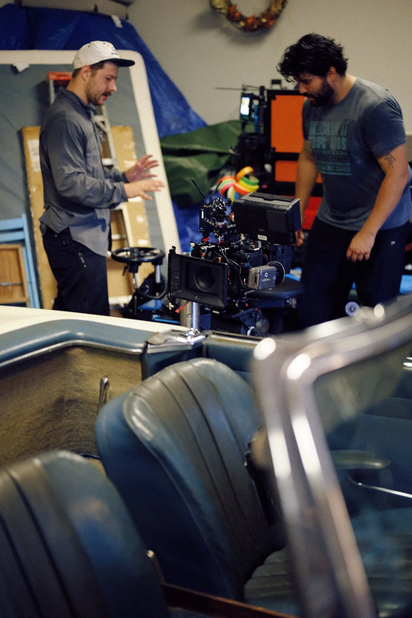 Two men on a film set working with a professional camera in a garage-like space, with vintage car seats in the foreground and a colorful wreath and storage items in the background.