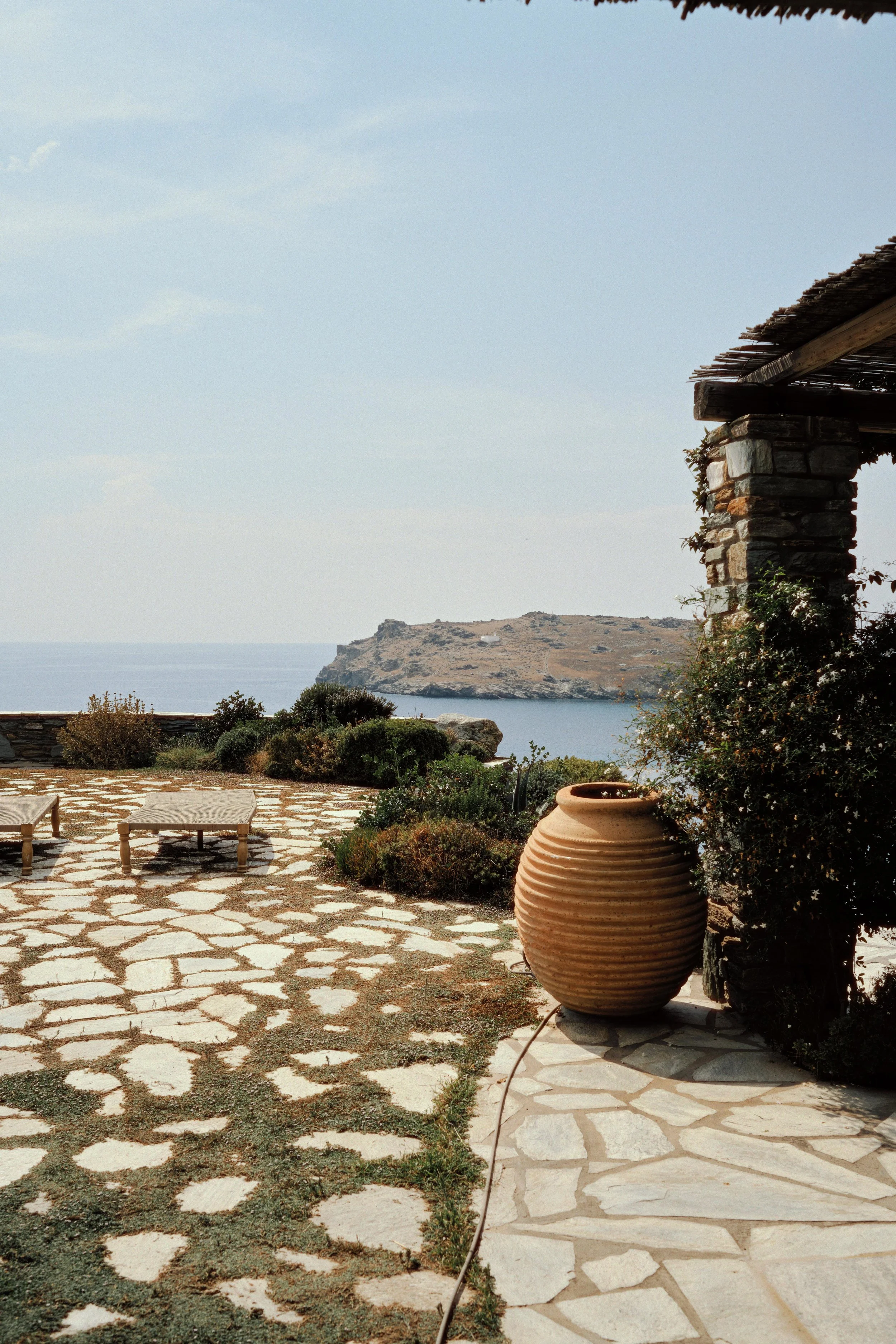 Outdoor patio with stone paving, two lounge chairs, large terracotta pot, bushes, and sea view with a hill in the distance under a partly cloudy sky.