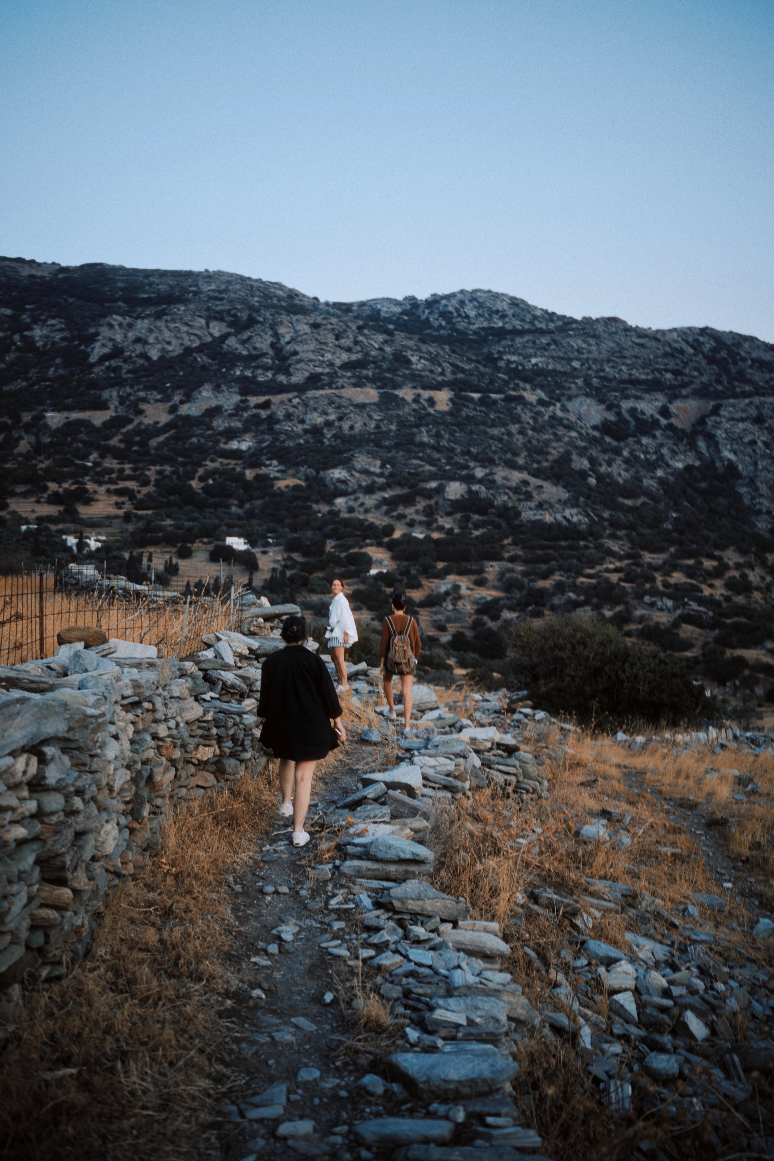 Three people walking on a rocky mountain trail during dusk with hills and sparse vegetation in the background.