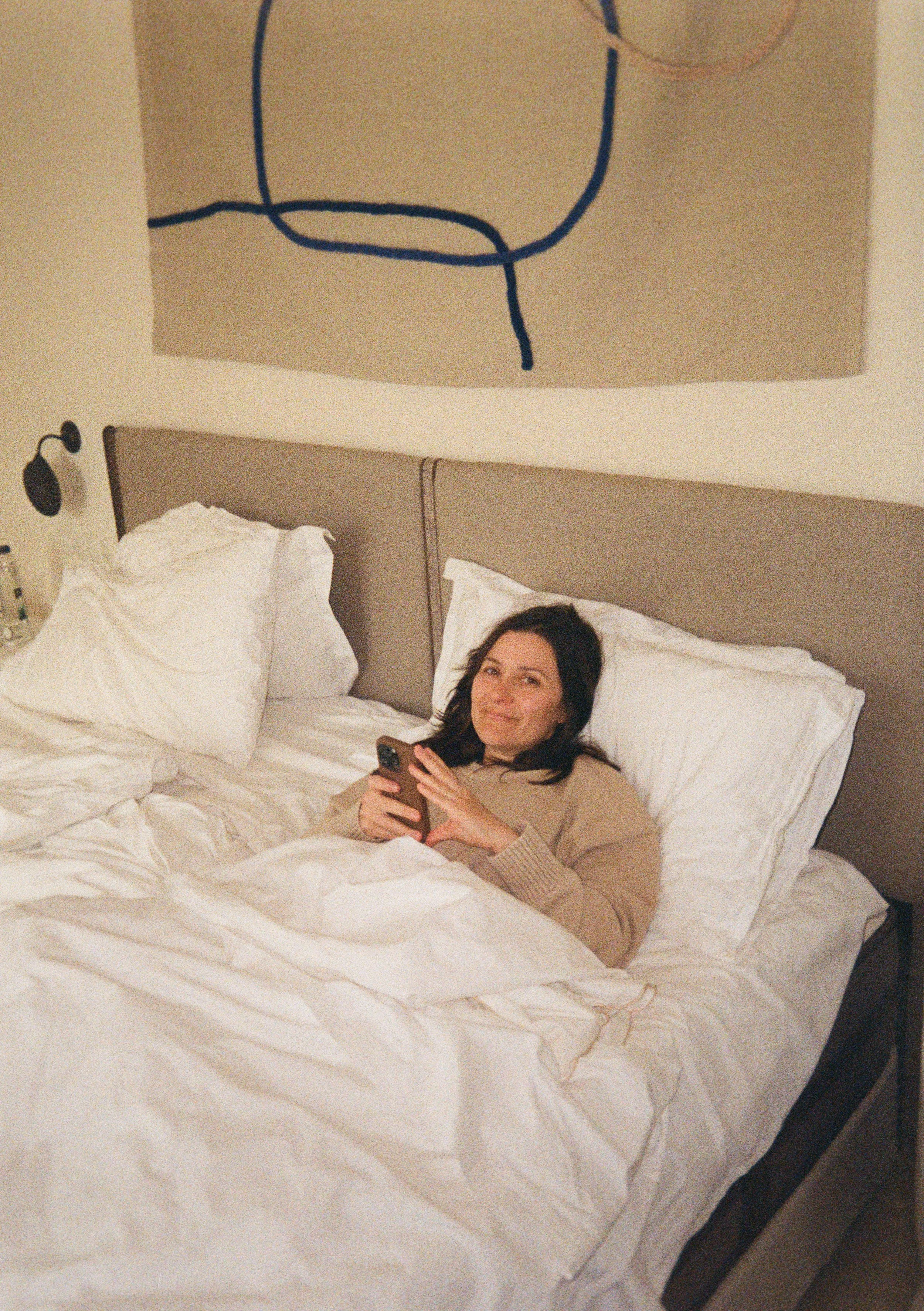 A woman with dark hair lying in bed, looking at the camera and holding a smartphone in her hands. The bed has white sheets and pillows, and there is abstract artwork hanging above the headboard.