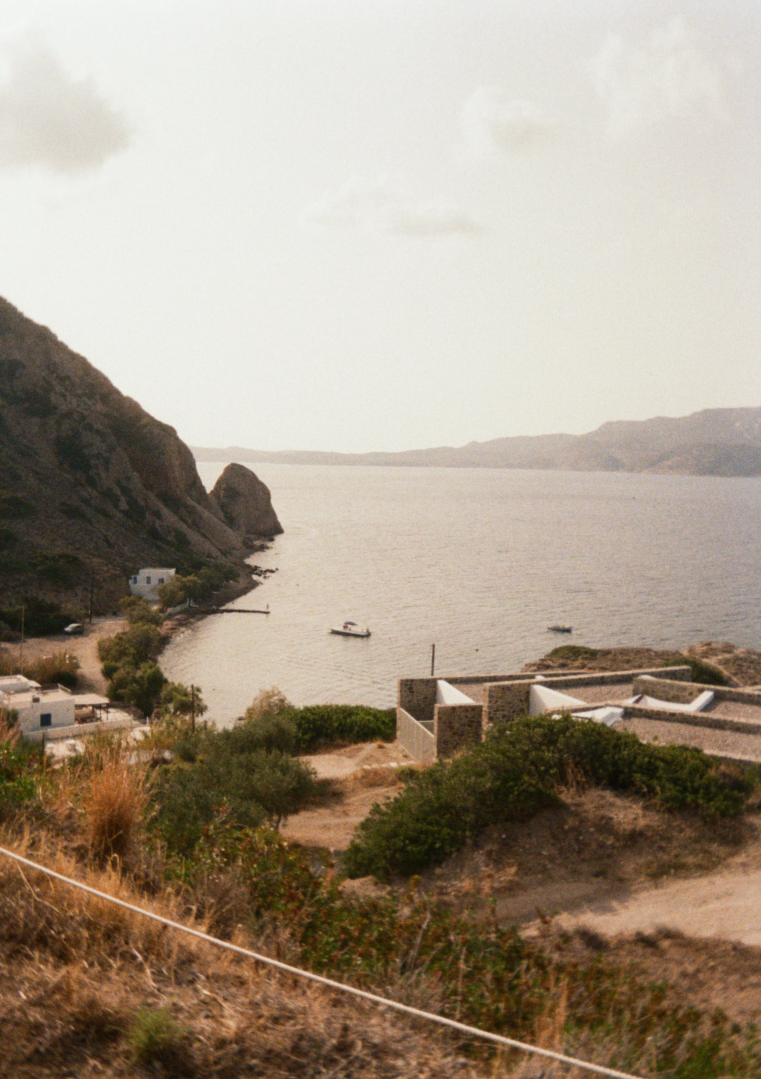 A coastal landscape with a calm ocean, a few boats floating, rocky hills, some small buildings, and sparse vegetation.