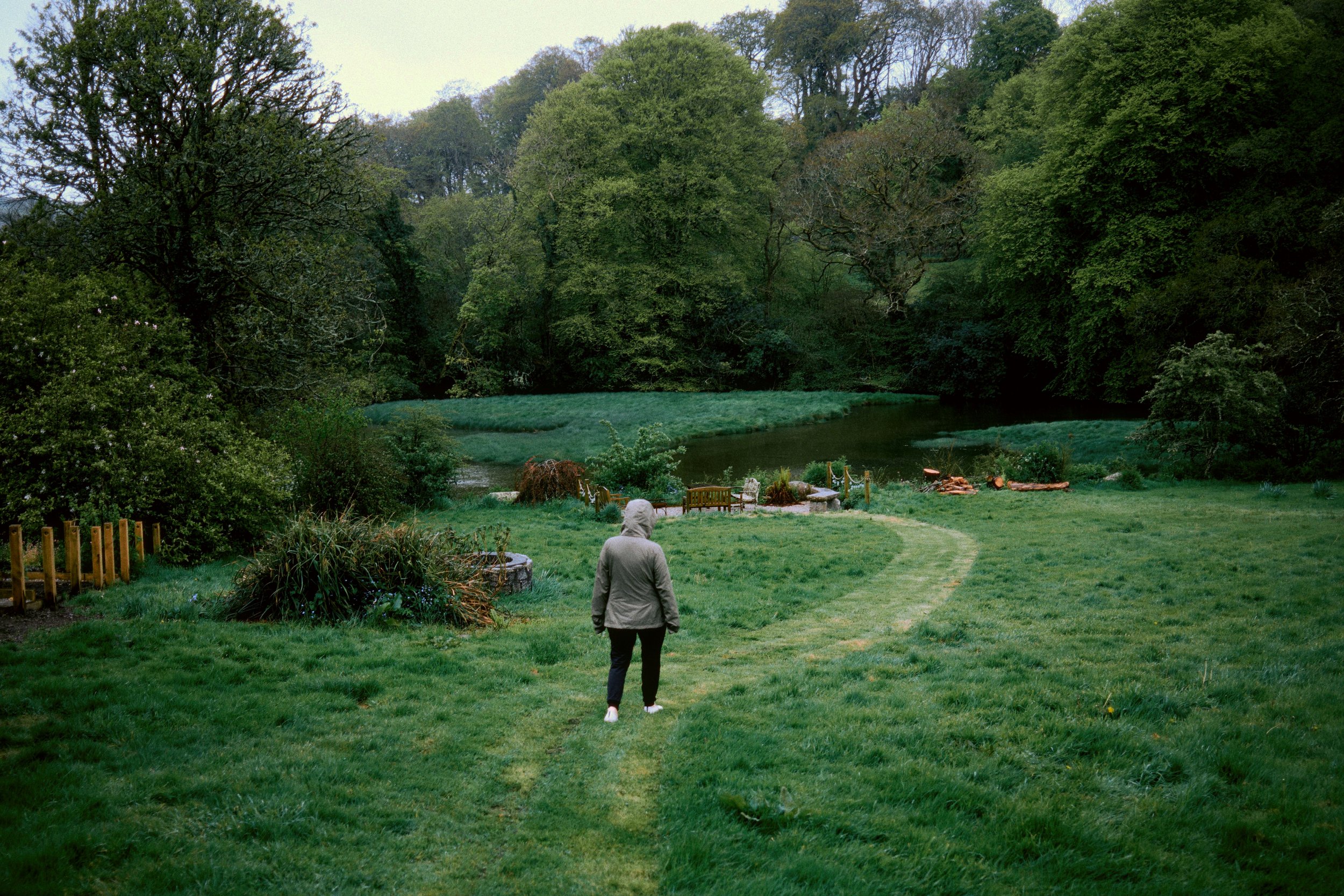 A person wearing a gray hoodie and black pants walking on a grassy path near a pond, surrounded by trees and greenery.