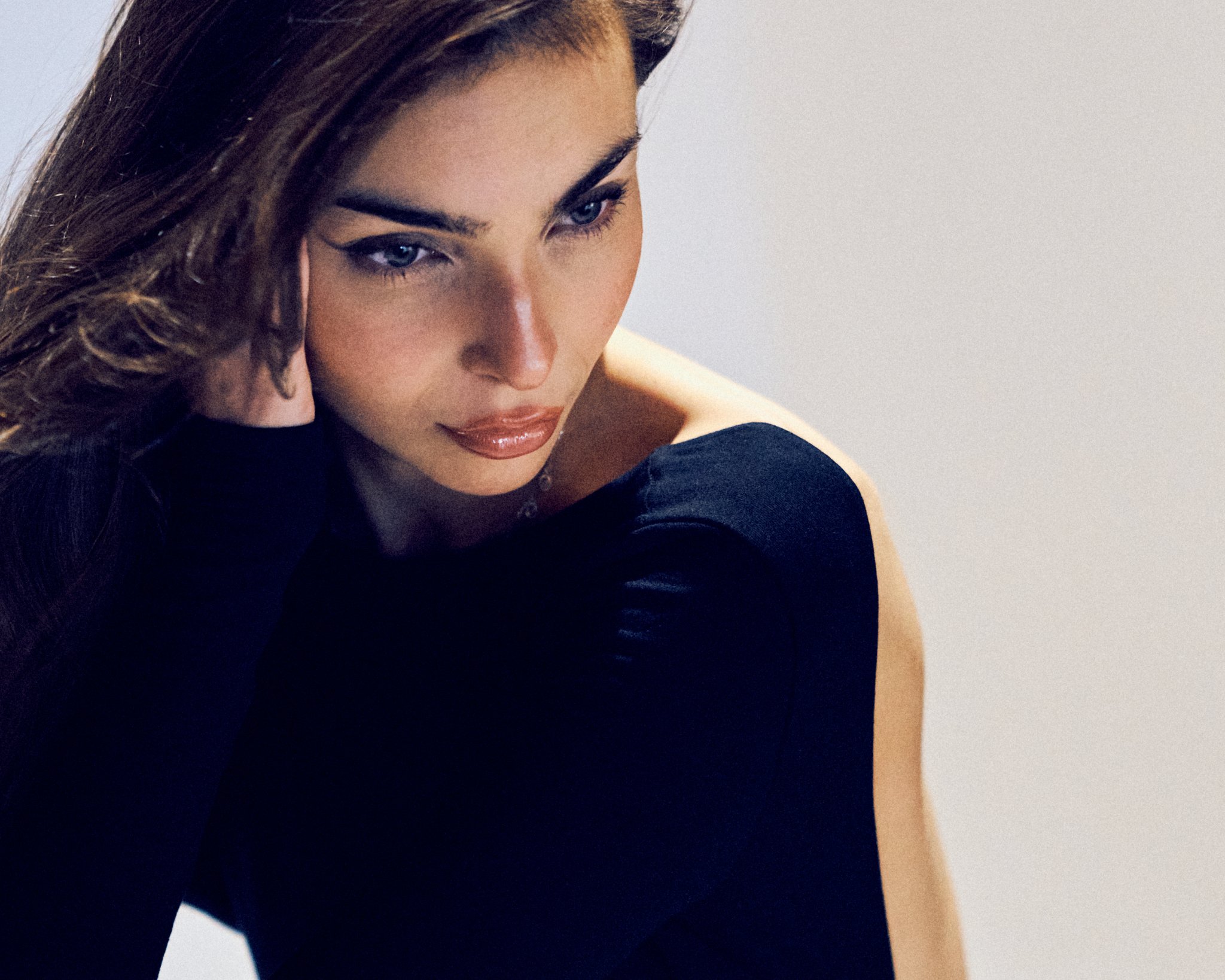 A woman with long, wavy brown hair and blue eyes poses with her hand near her face, wearing a black long-sleeve top against a plain light background.