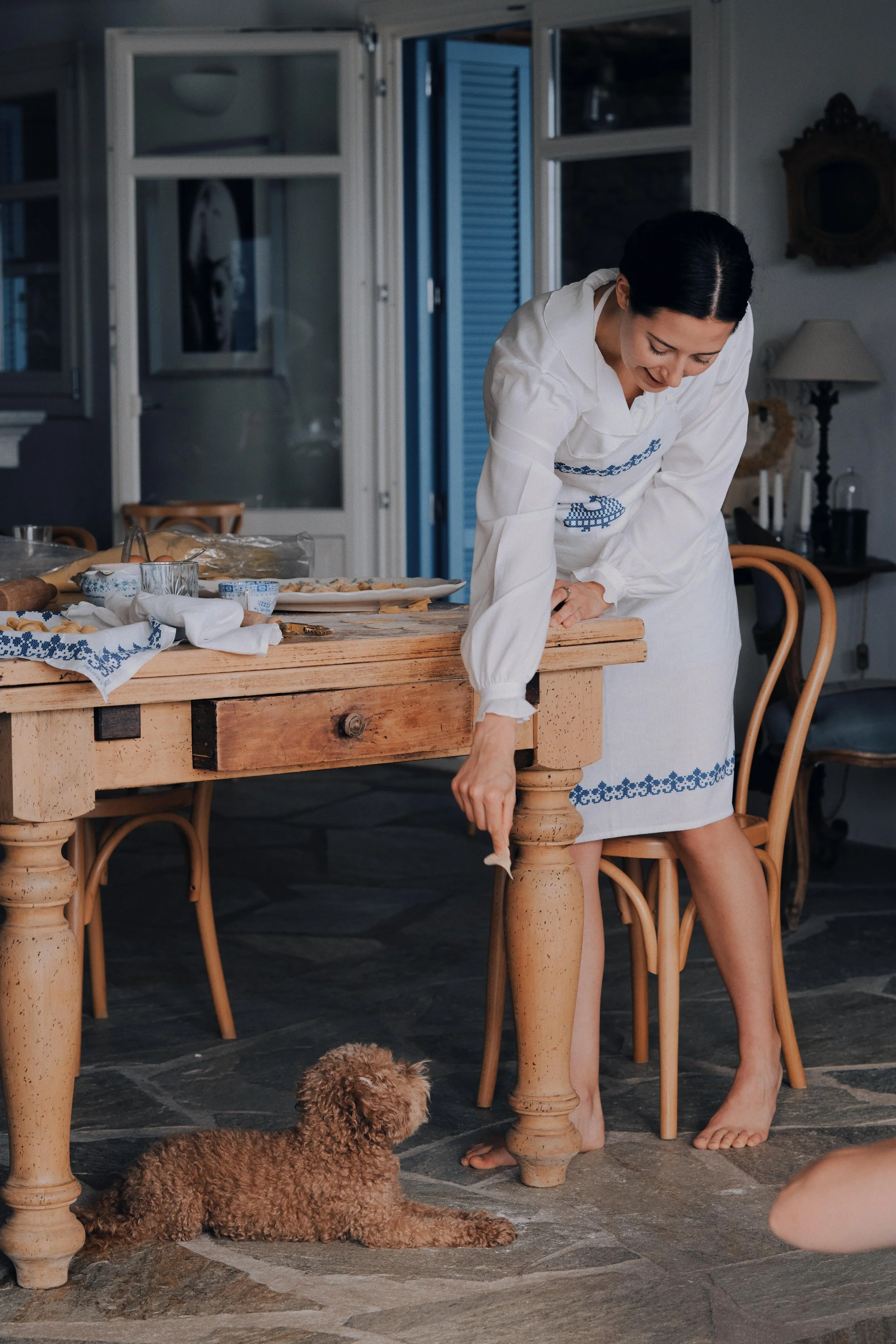 A woman in a white dress with blue embroidery cleaning the floor underneath a wooden dining table, with a small brown poodle lying nearby in a cozy, decorated kitchen or dining area.