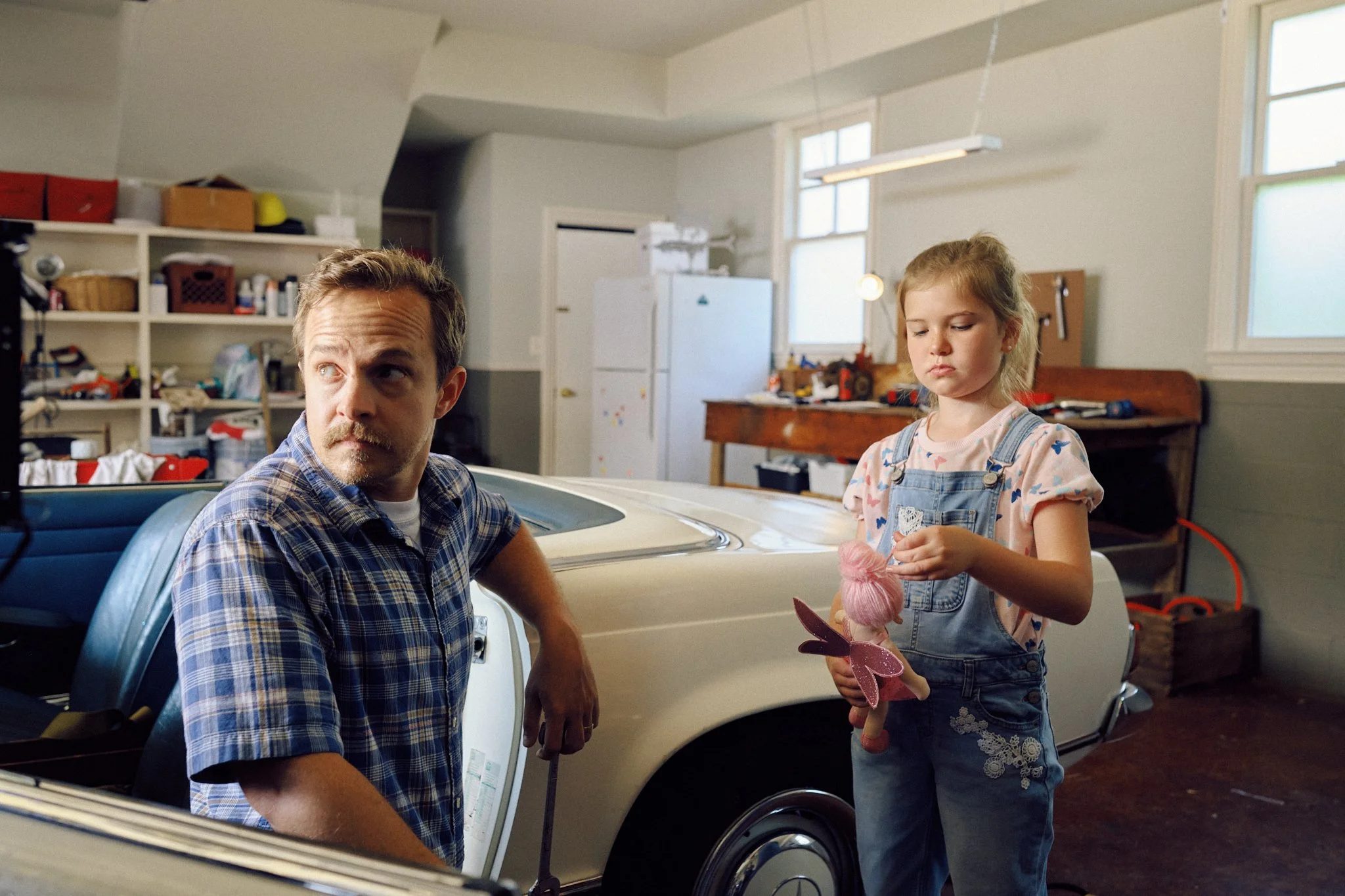 A man and a young girl in a garage with a vintage car; the man is sitting, looking to the side, while the girl stands holding a pink stuffed animal and looking downward.
