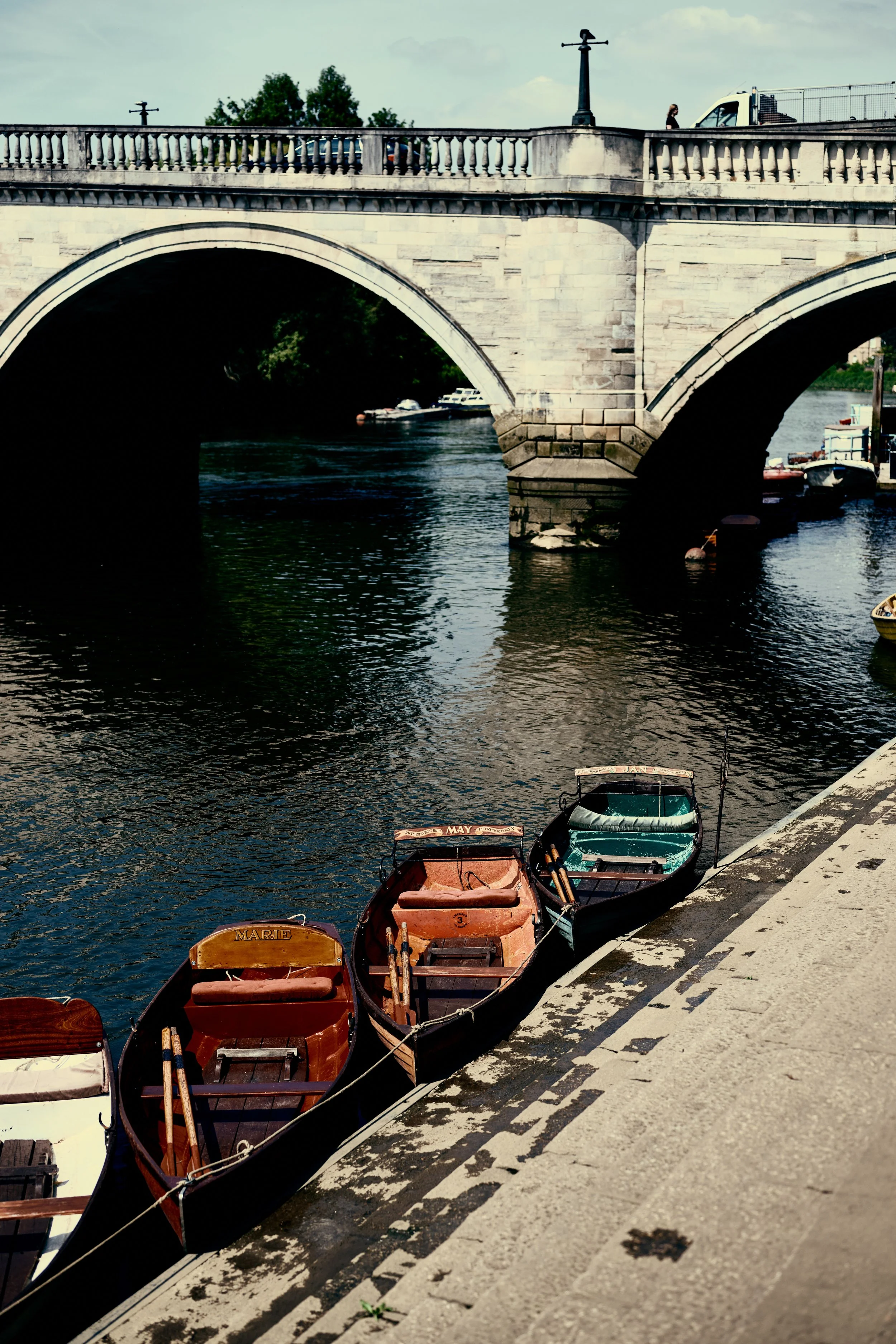 A stone bridge over a river with boats docked along the riverbank in a city setting.