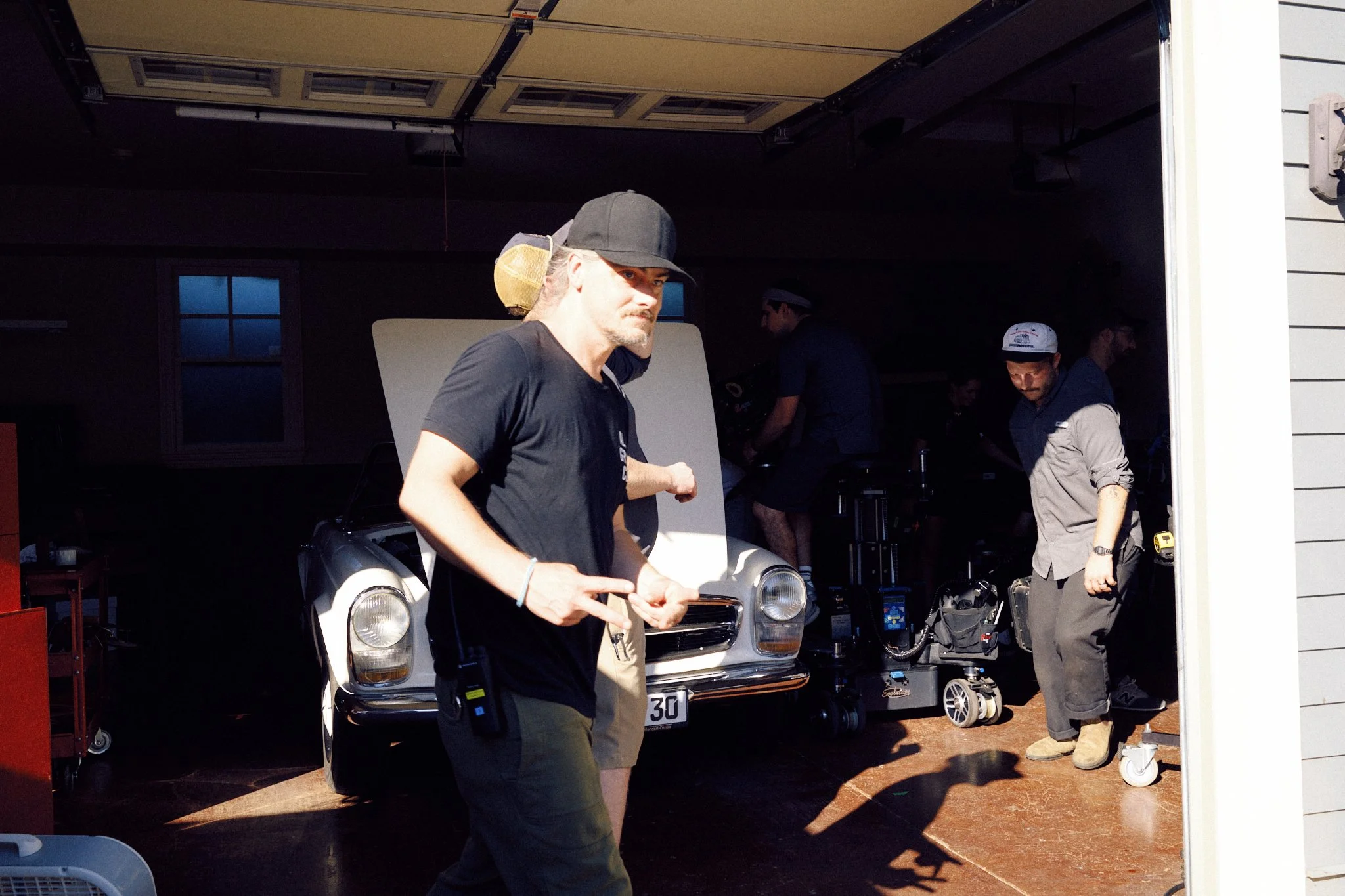 A man with long hair and a beard walking out of a garage, wearing a black cap and black T-shirt. Inside the garage, several people are working with filming equipment and a vintage car with the hood open.