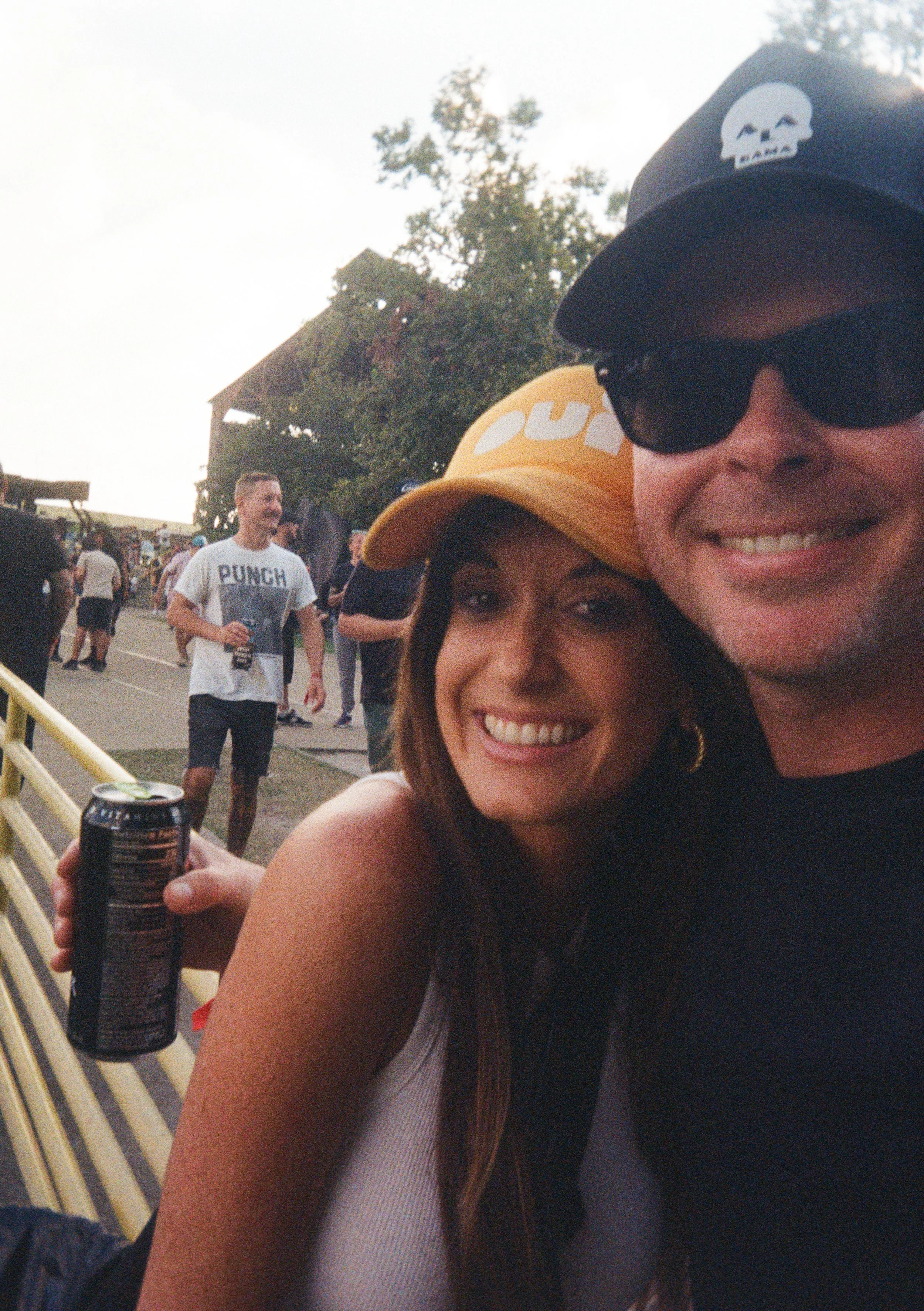 A man and woman taking a selfie at an outdoor event, both wearing branded baseball caps and smiling. There are other people and trees in the background, with one person holding a drink.