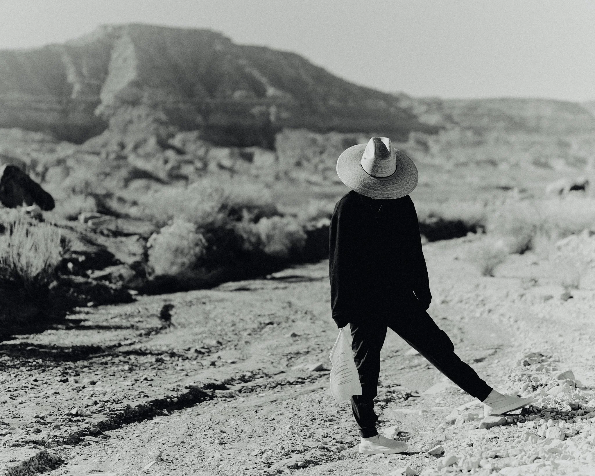 A person wearing a wide-brimmed hat, dark clothing, and sneakers walking on a rocky desert trail, with a rocky hill in the background.
