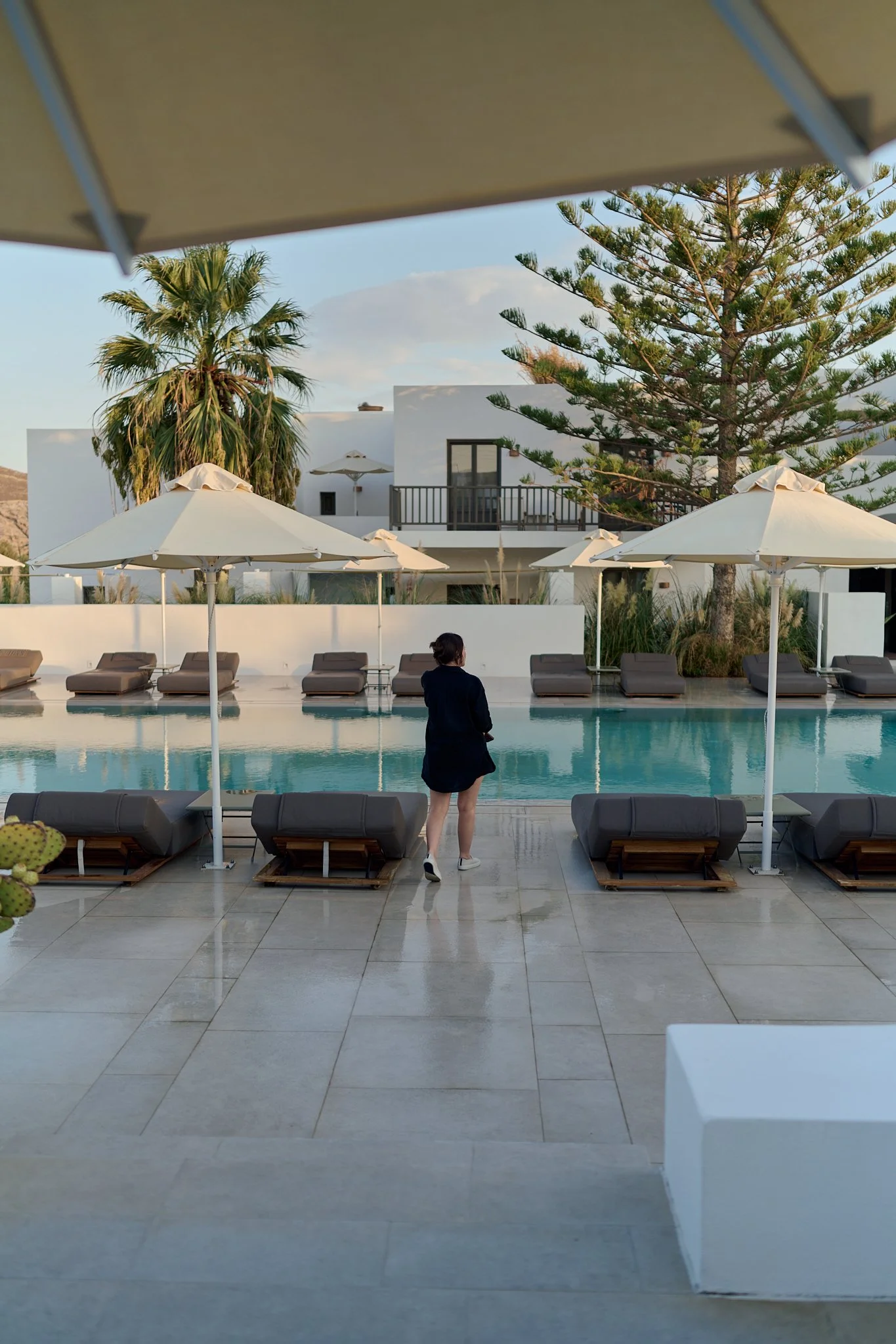 View of a swimming pool area with lounge chairs, umbrellas, a person walking, and modern white buildings in the background, surrounded by trees.