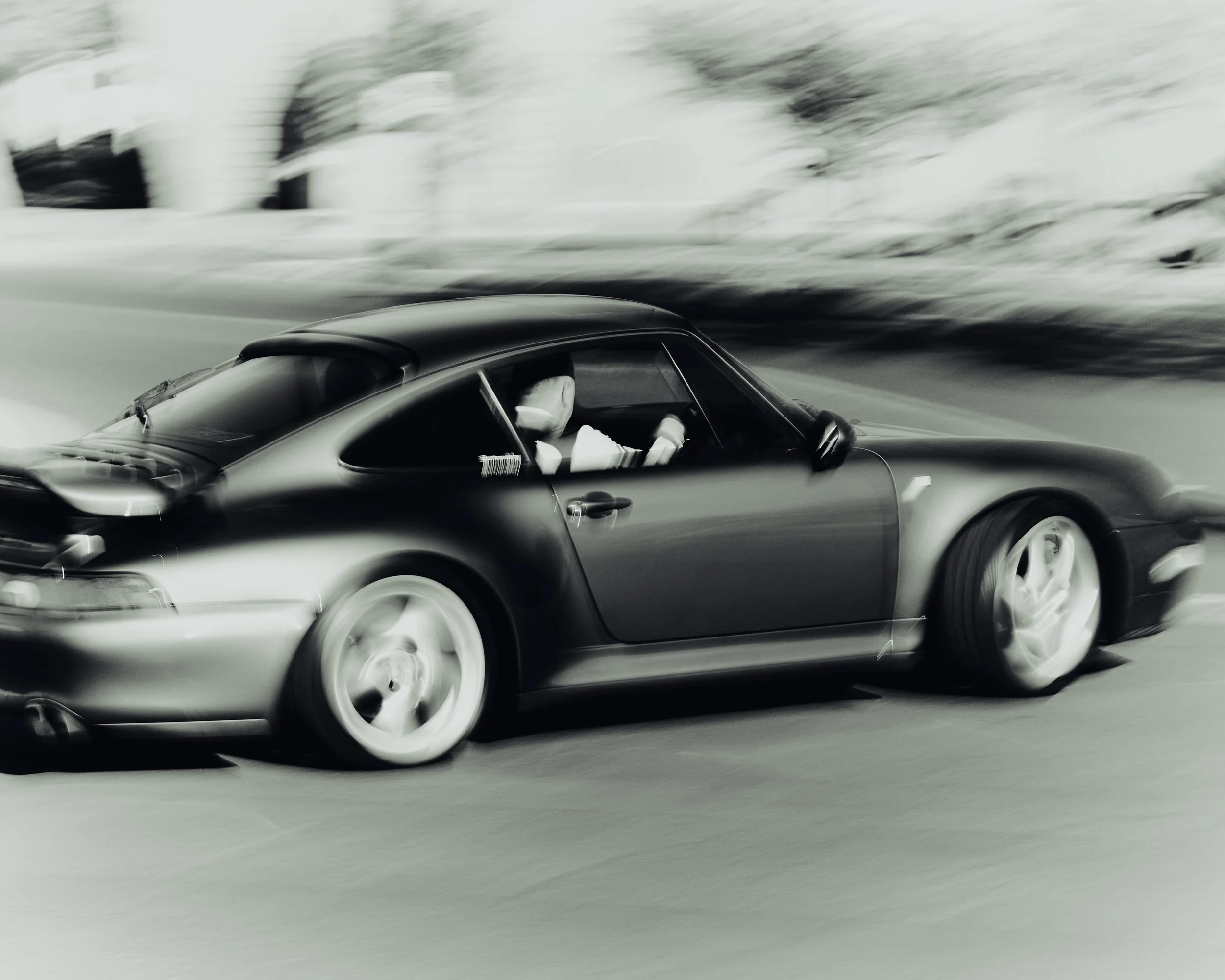 Black and white photo of a sports car racing on a track, with a driver visible inside, motion blur indicating high speed.