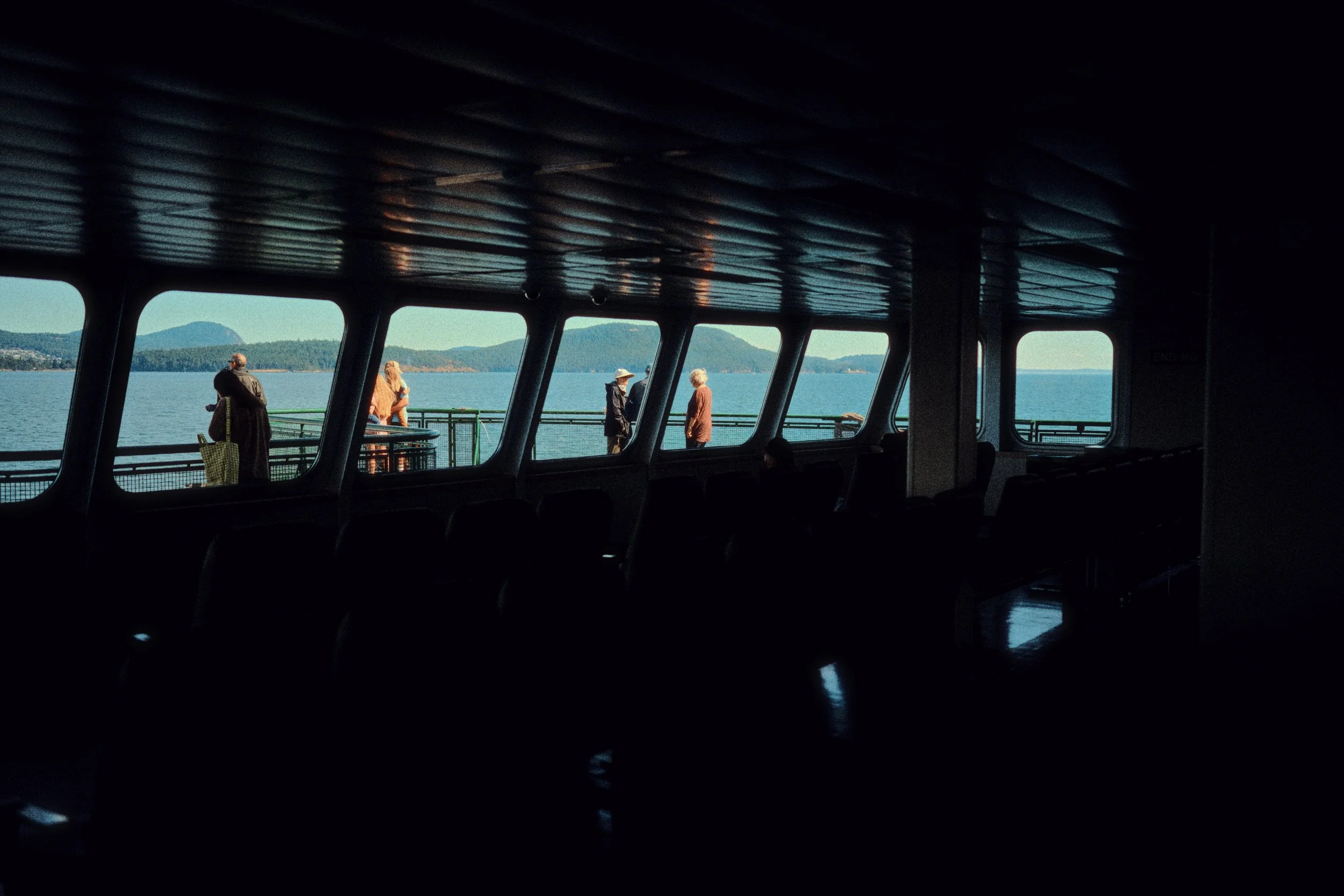 People standing outside on a ferry deck, looking at the water and mountains.