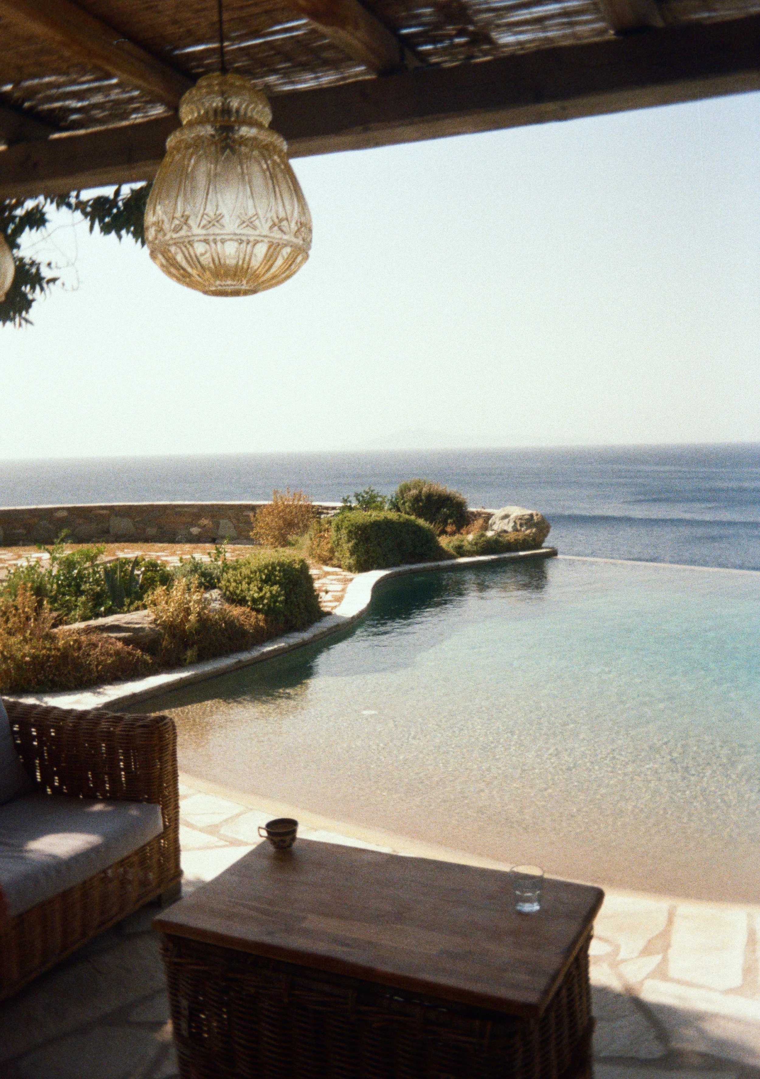 Outdoor patio with wicker furniture, a small coffee table, and a pool overlooking the ocean under a wooden roof with a hanging light fixture.