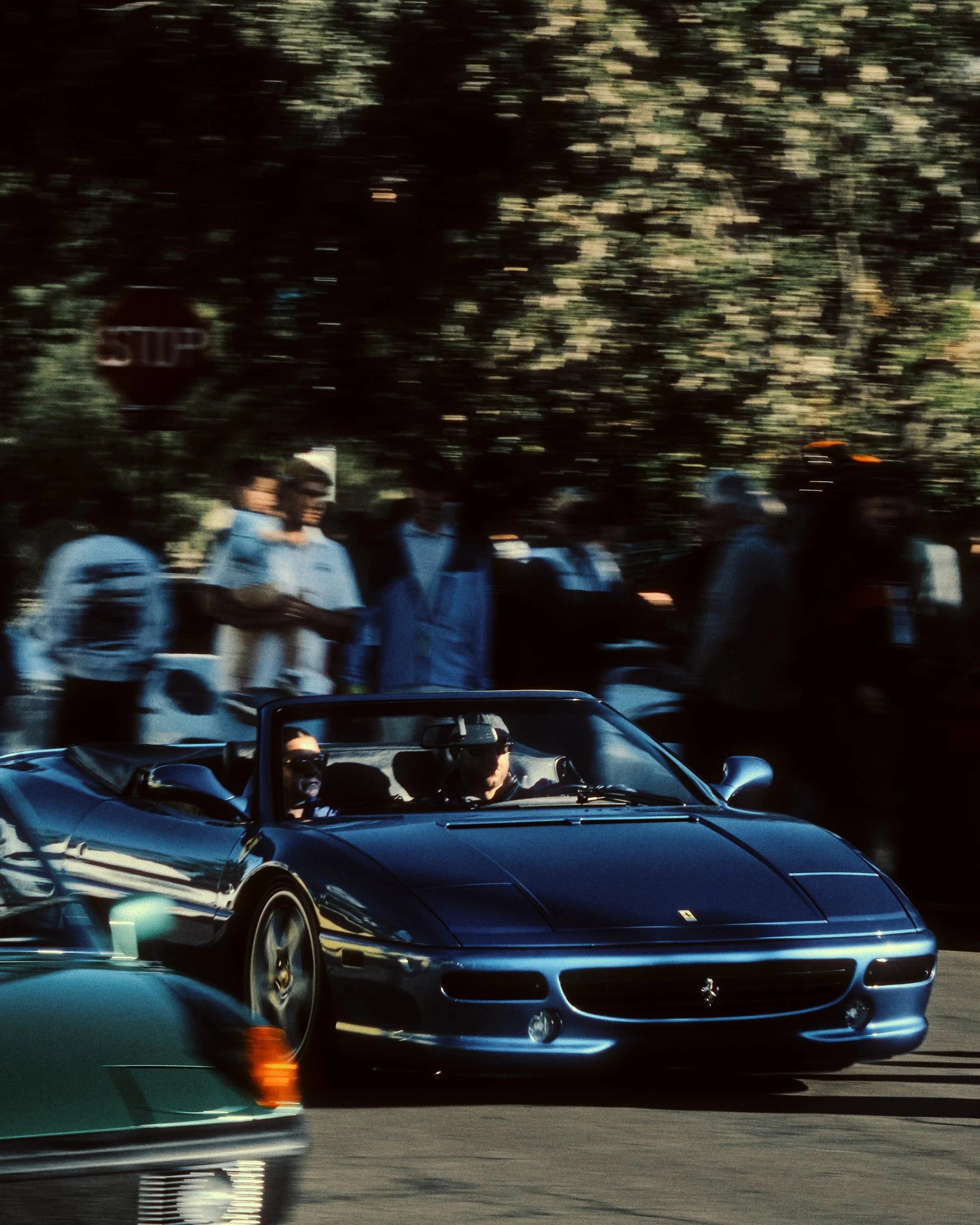A black convertible Ferrari driving on a street with a blurred background of pedestrians walking, some wearing sunglasses and hats, and trees.
