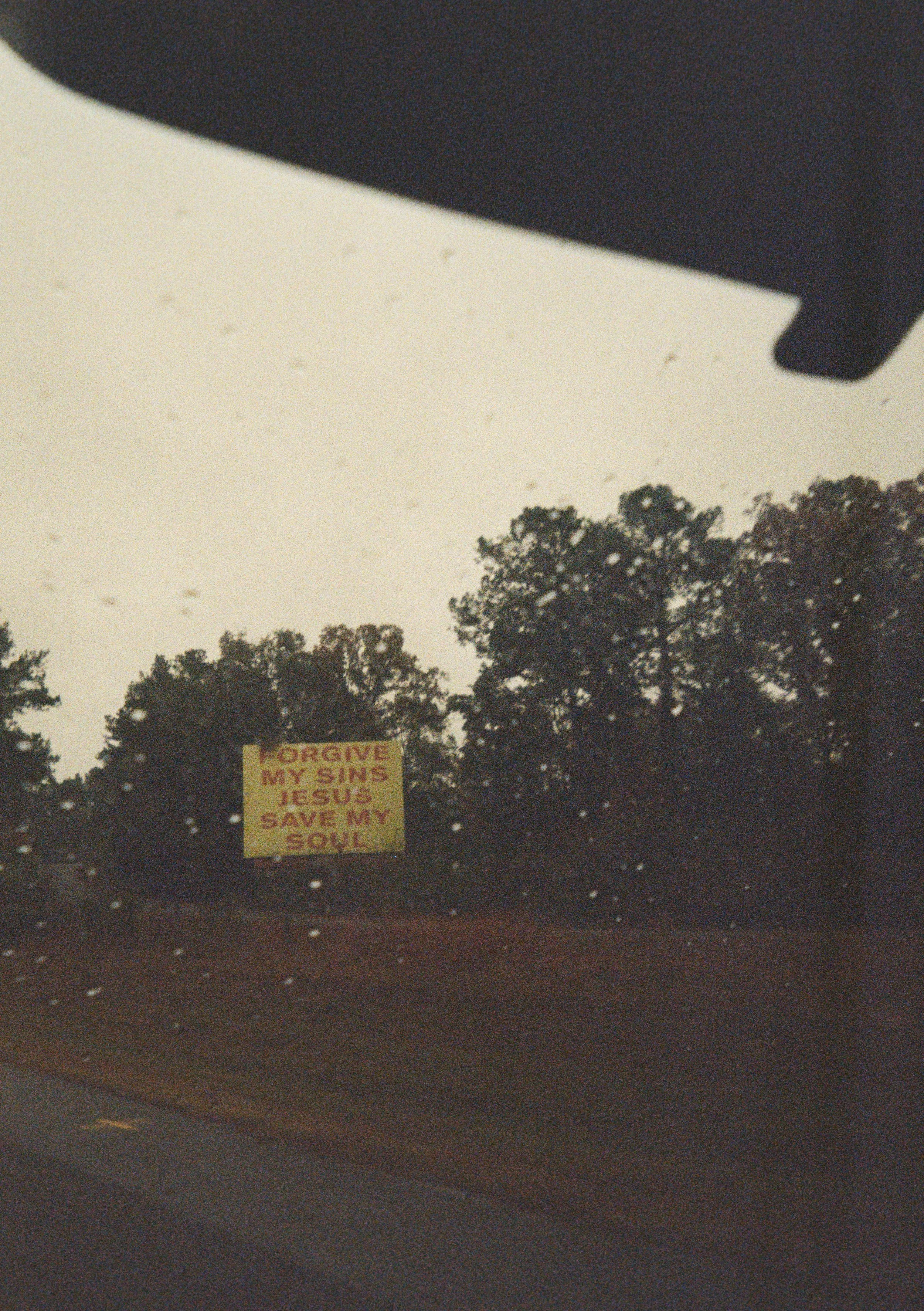A yellow sign with red text that reads, 'FORGIVE MY SINS JESUS SAVE MY SOUL' visible in the distance against trees and a cloudy sky on a rainy day.