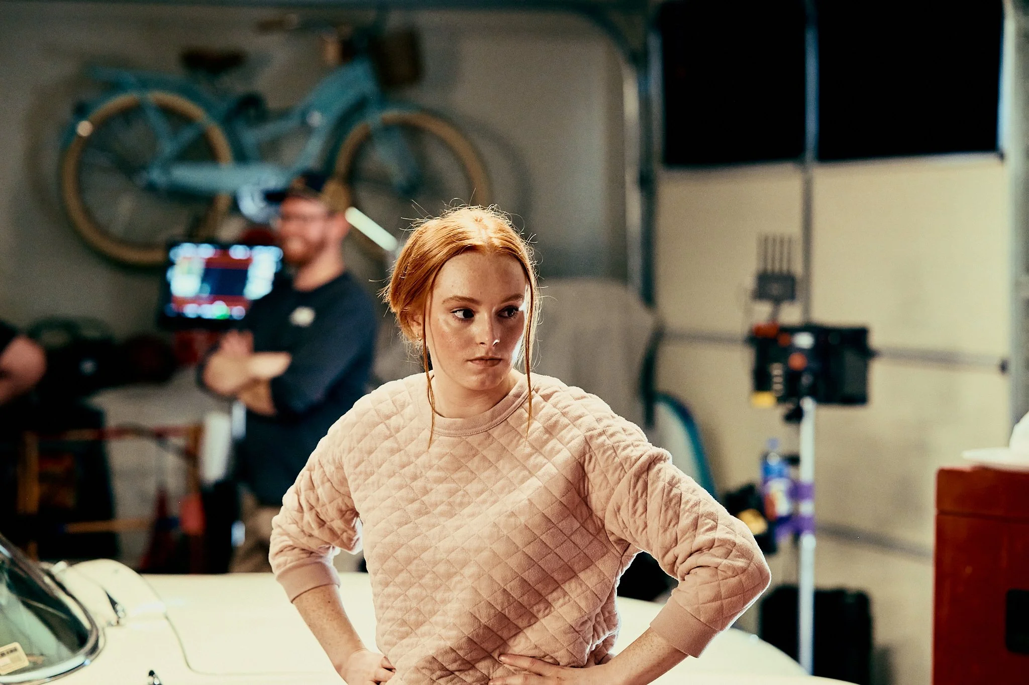 A young woman with red hair and a pink quilted sweatshirt, standing with her hands on her hips in a garage, with a woman in the background blurred, near a blue bicycle mounted on the wall.