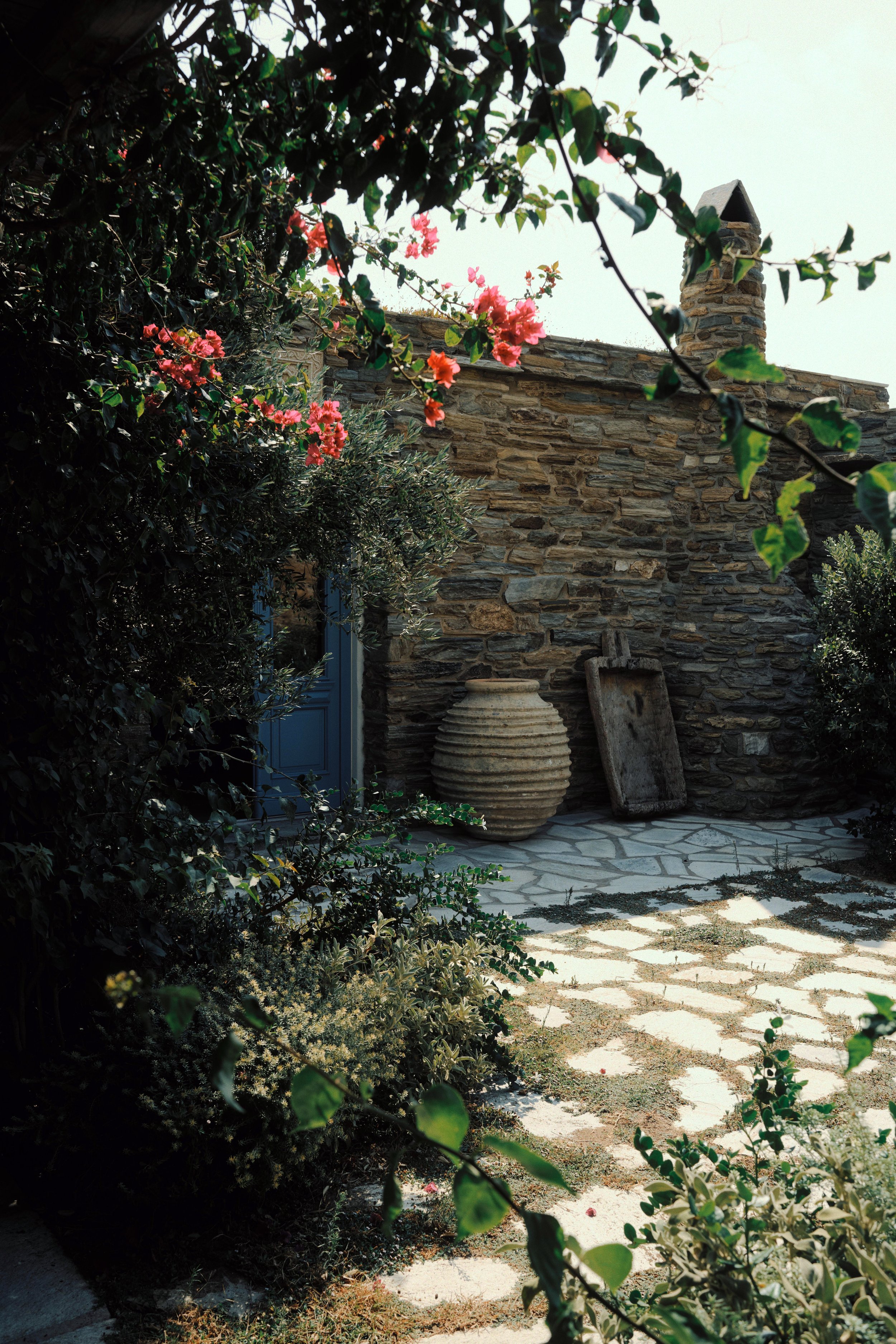 A stone patio with a large round woven clay pot and a worn wooden tray leaning against a stone wall surrounded by lush green plants and pink flowering bushes.
