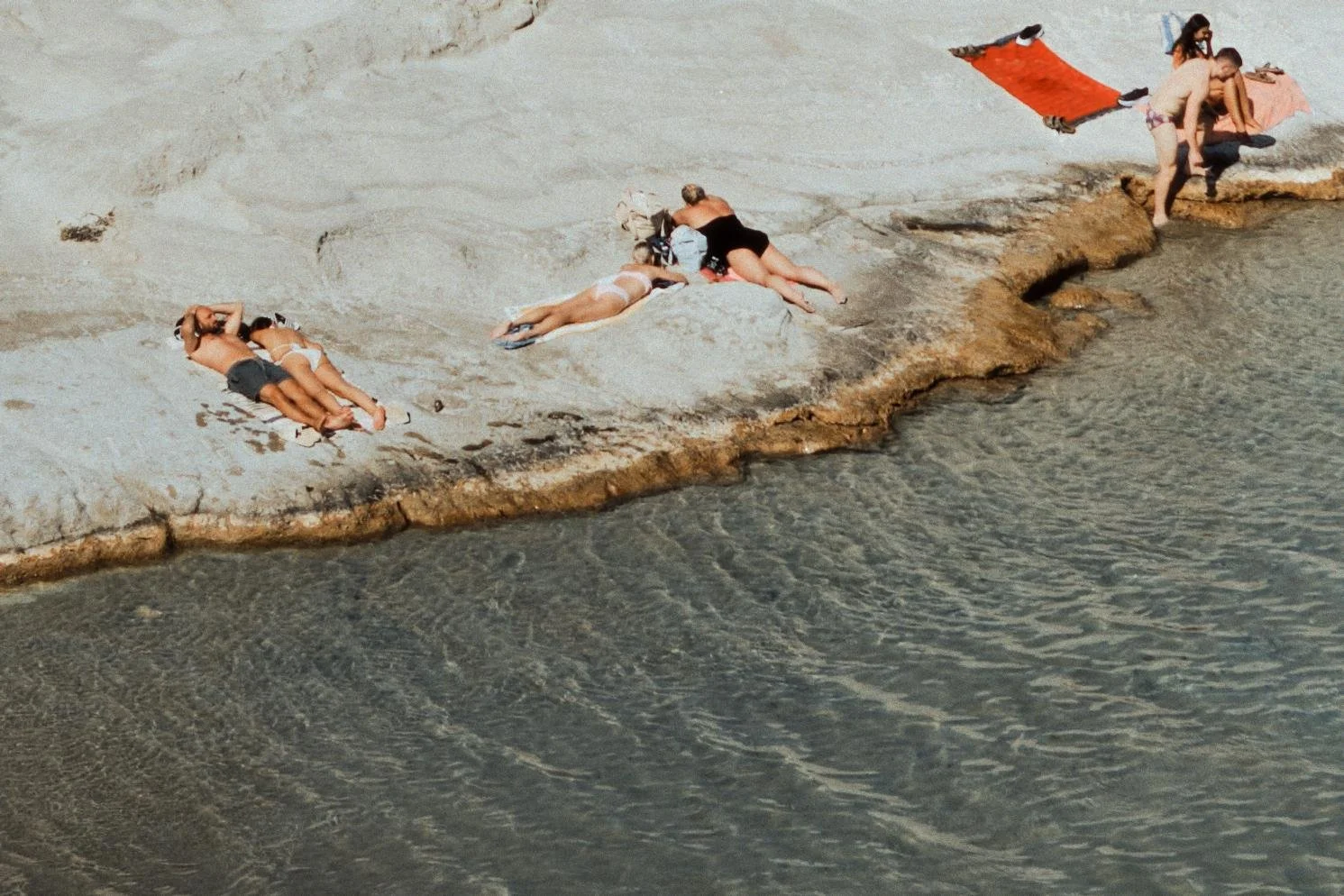 People sunbathing on a rocky shoreline near the water, with some lying on towels and others sitting or standing nearby.