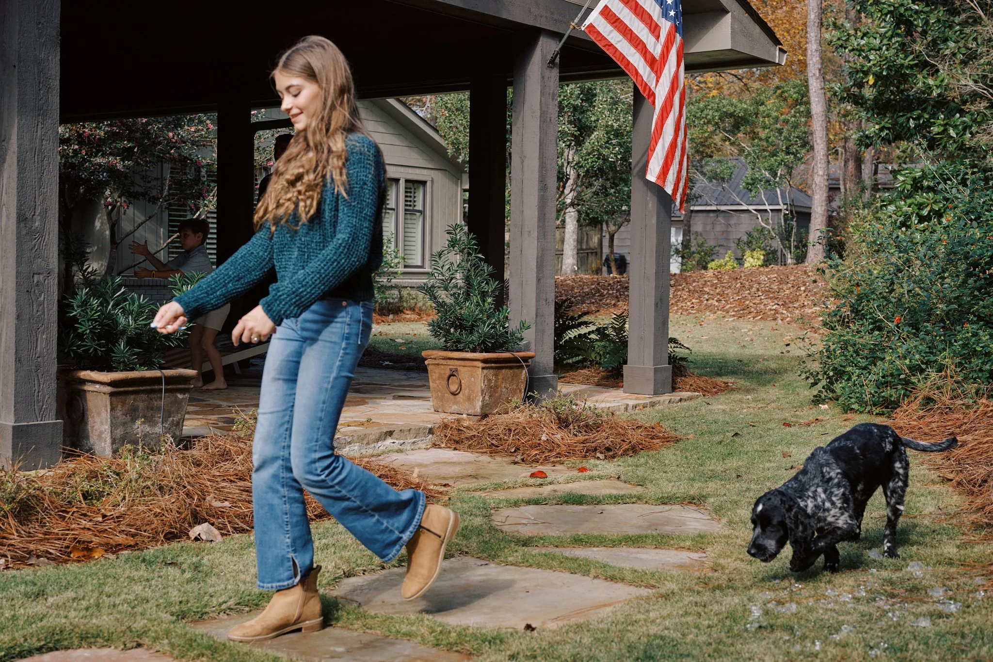 A girl with long hair in a teal sweater and jeans is playing outdoors in front of a house with a flag, while a black and white puppy walks nearby. A young boy is sitting on a porch in the background.