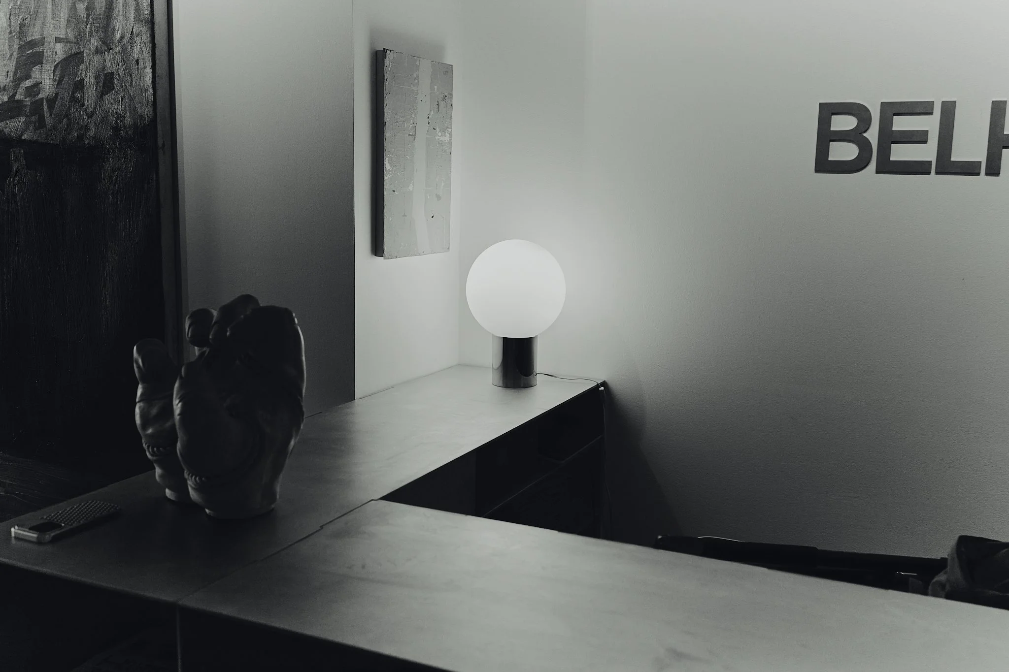 A minimalist indoor scene featuring a white textured wall with partial black text, a wooden cabinet with a glowing table lamp, two decorative handles resembling hands, a remote control, and a painting on the wall.