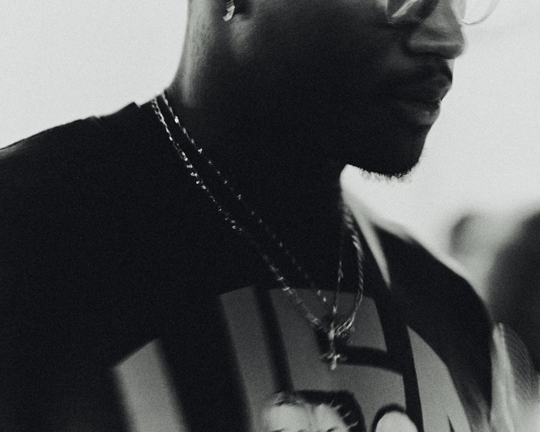 Close-up black and white photo of a person's face and upper body, wearing glasses, necklaces, and a graphic t-shirt.