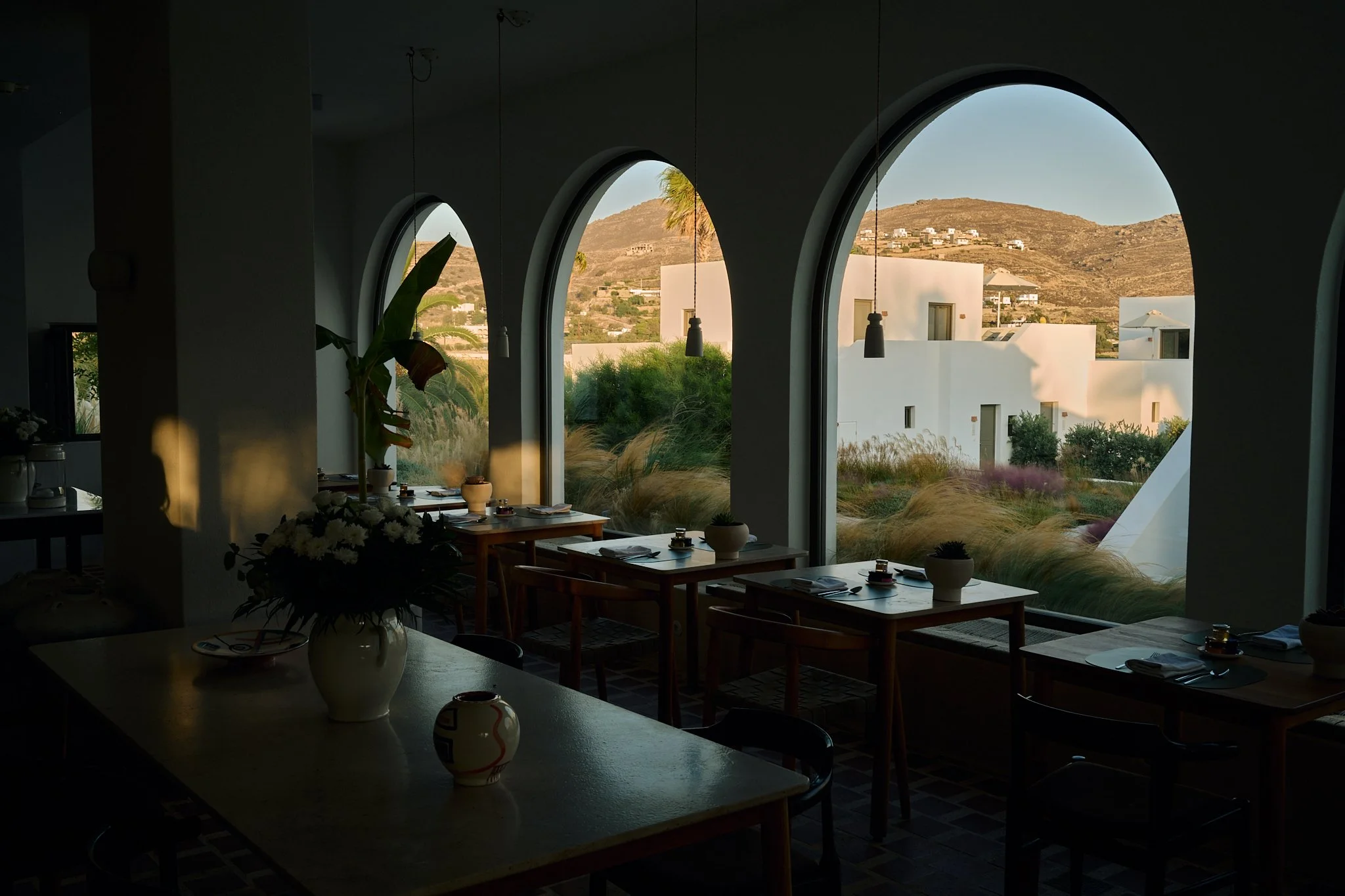 Interior of a restaurant with large arched windows showing a view of hills and modern white buildings outside. The restaurant has tables set with plates, cups, and small potted plants, and there are hanging pendant lights.
