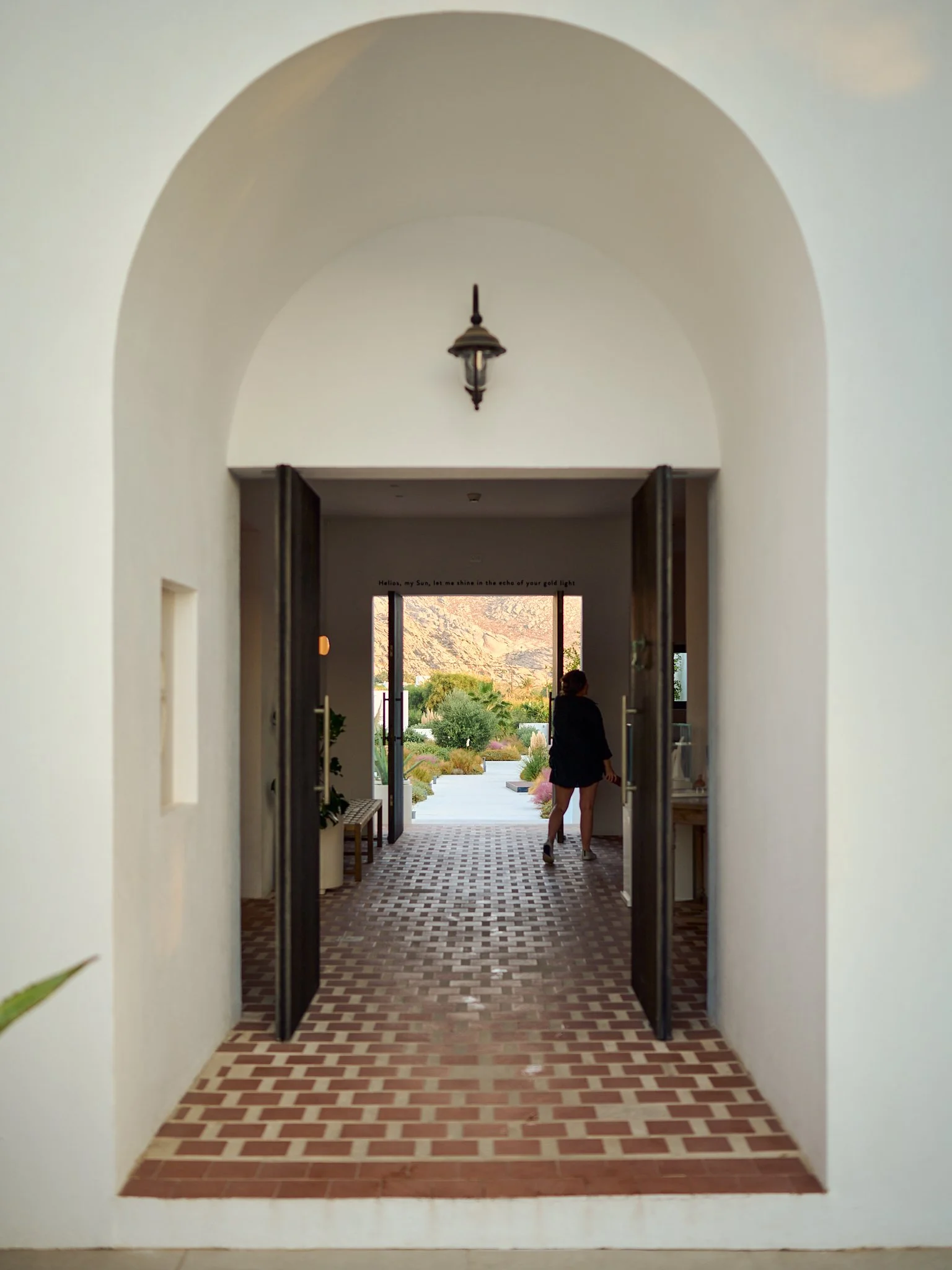 View through an open doorway, showing a person with curly hair in a black outfit walking towards a sunlit outdoor landscape with mountains in the background.