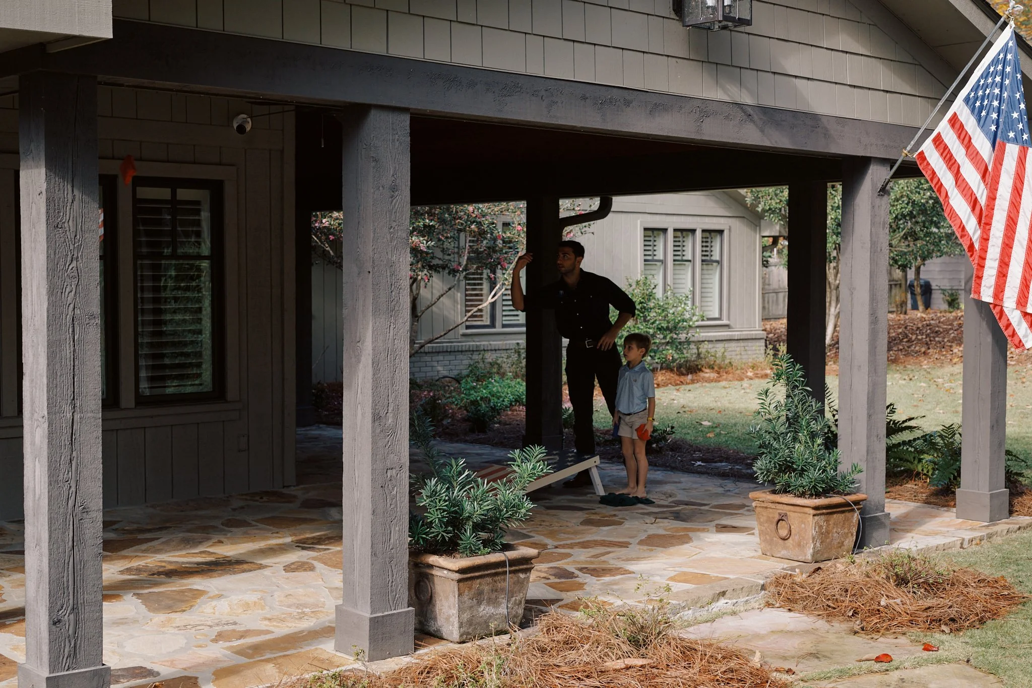 A man and a young boy with a toy watering can on a porch with stone flooring, potted plants, and an American flag.