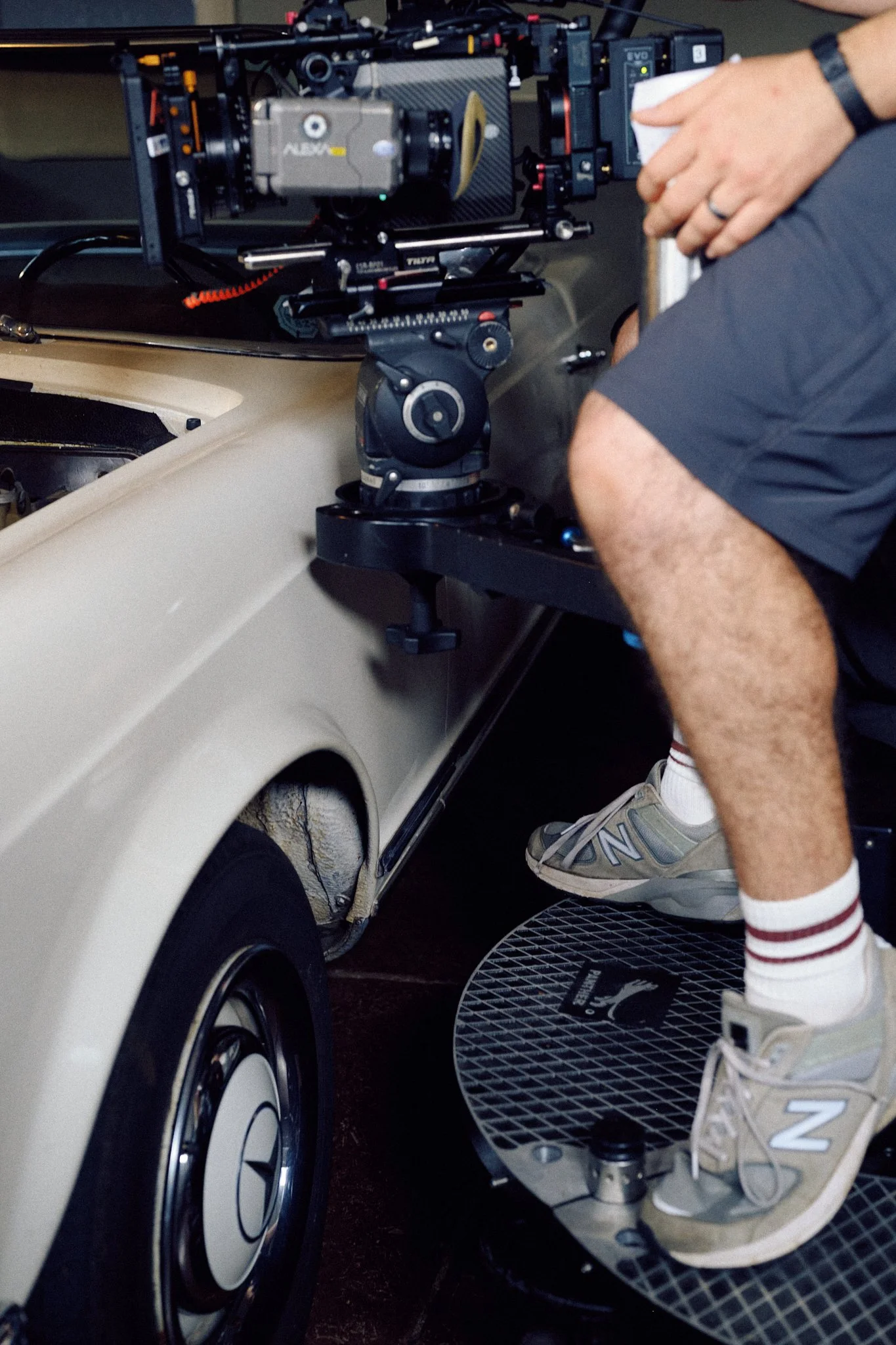 Close-up of a person operating a camera mounted on a tripod attached to a vintage car, with the person's legs and sneakers visible, along with a Puma-branded workout disc beneath his feet.