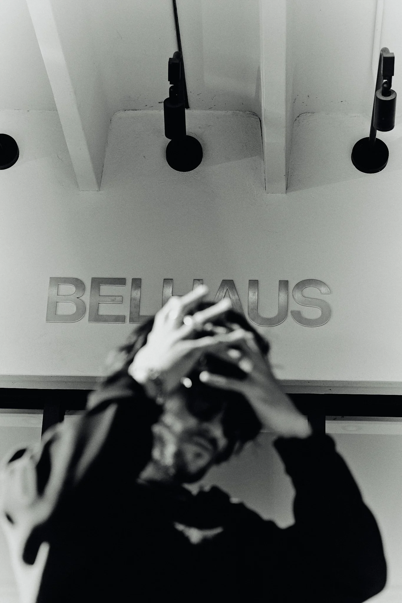 Black and white photo of a person with glasses holding their head, looking up towards a ceiling with the word "BELKAUS" on it.